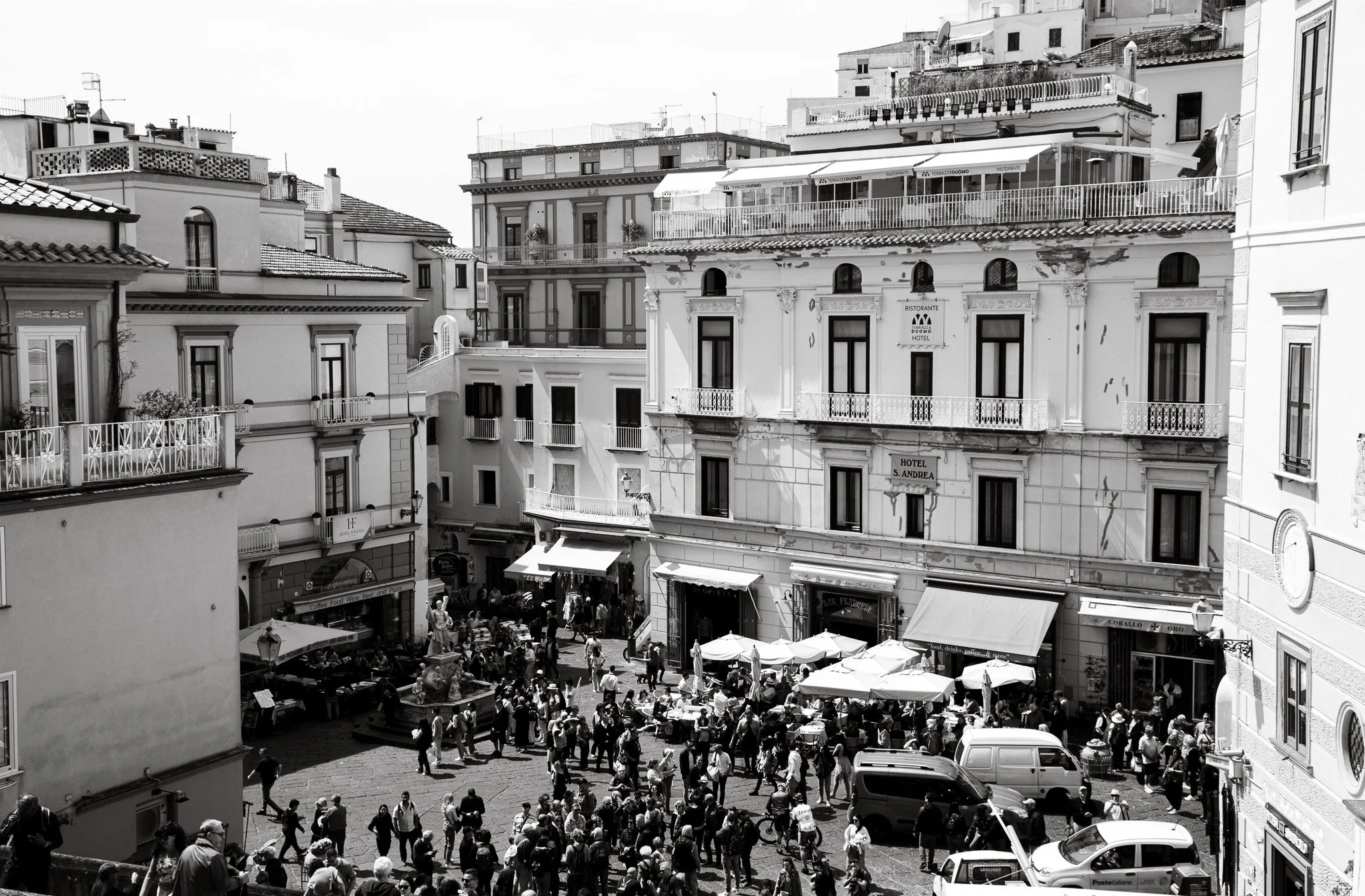 Black and white photo of a busy outdoor market or town square with many people, umbrellas, and multi-story buildings with store signs and balconies.