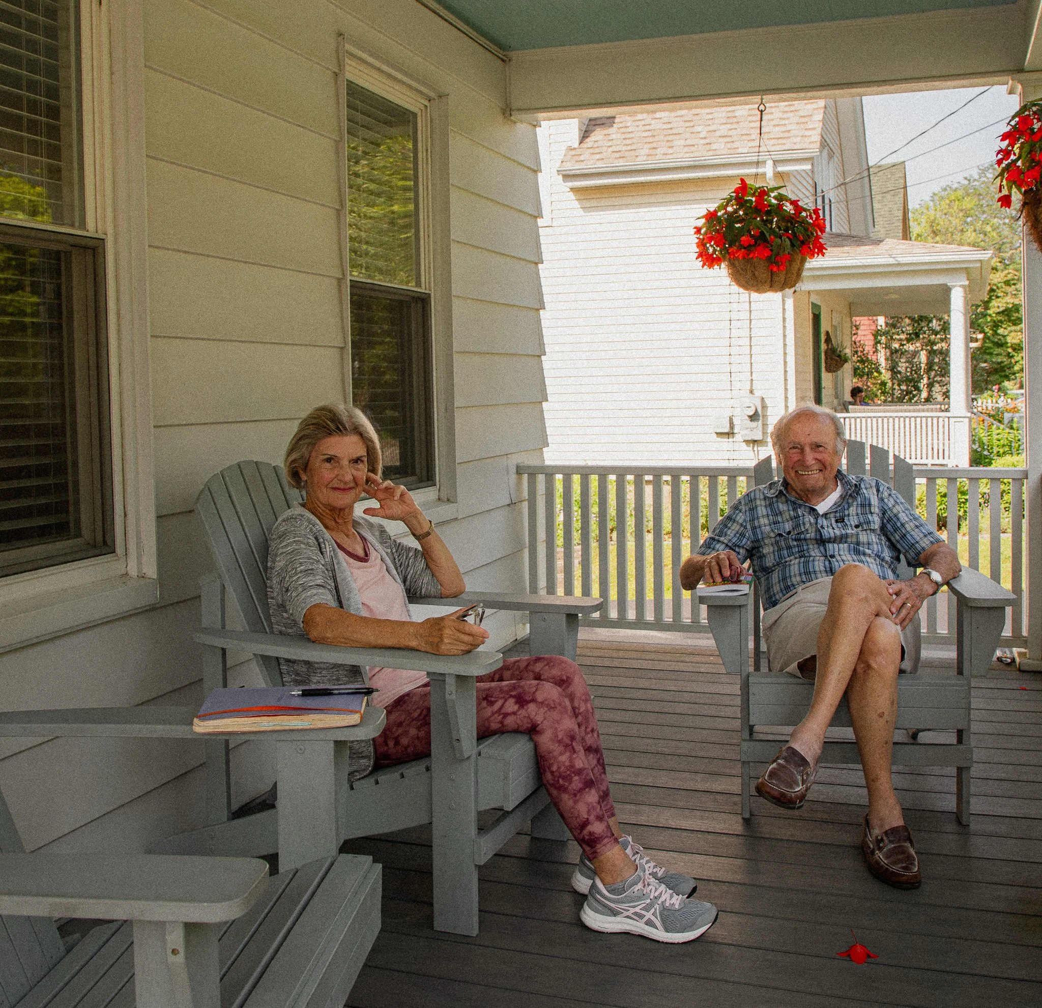 Two older adults, a woman and a man, sitting on a porch with light-colored siding, a window, hanging flower baskets, and outdoor furniture. The woman wears casual clothes and sneakers, resting her chin on her hand, while the man wears a plaid shirt a