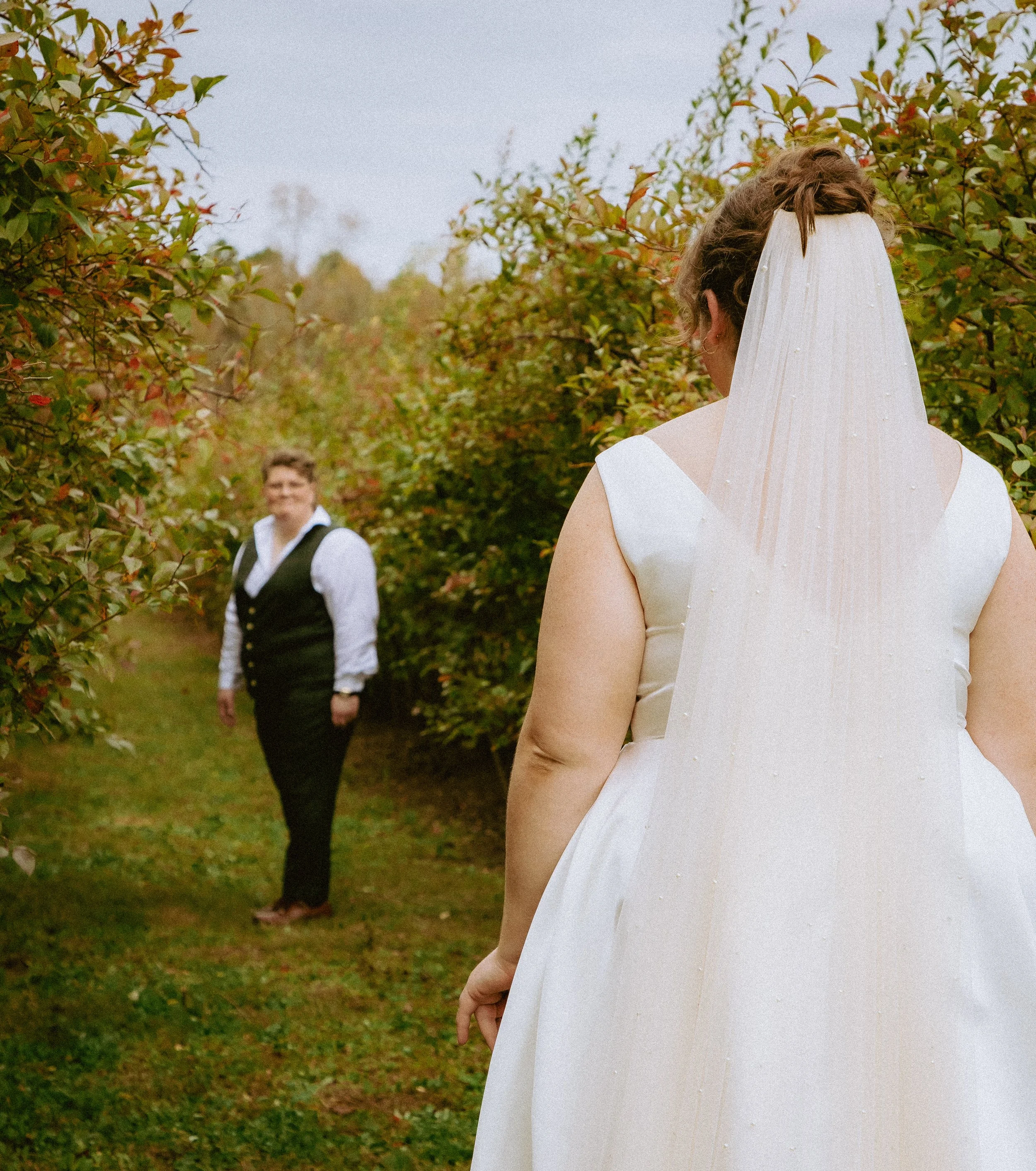 A bride in a white wedding dress and veil stands outdoors among trees, with a person in a white shirt and black vest in the background.