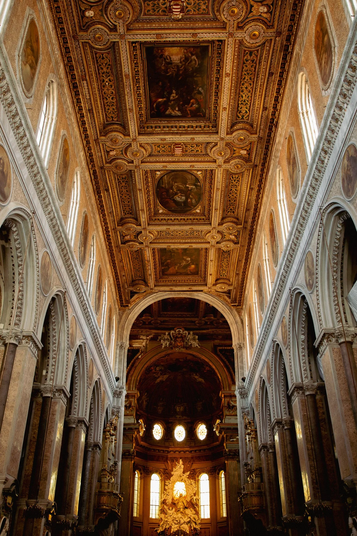 Interior of a grand, ornately decorated church or palace with a high, elaborately painted ceiling, tall arched windows, and a large altar featuring a bright white sculpture.