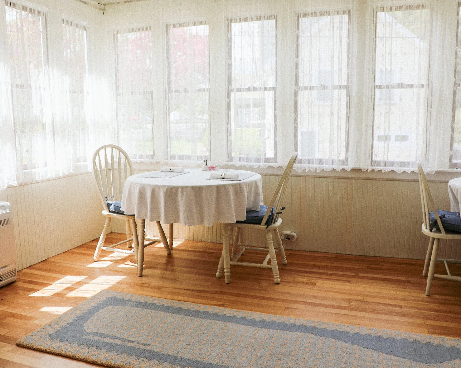 A dining area with a round table covered with a white tablecloth, set for two with napkins and utensils, surrounded by four wooden chairs with dark seat cushions, in front of large windows with sheer white curtains and hardwood floors.