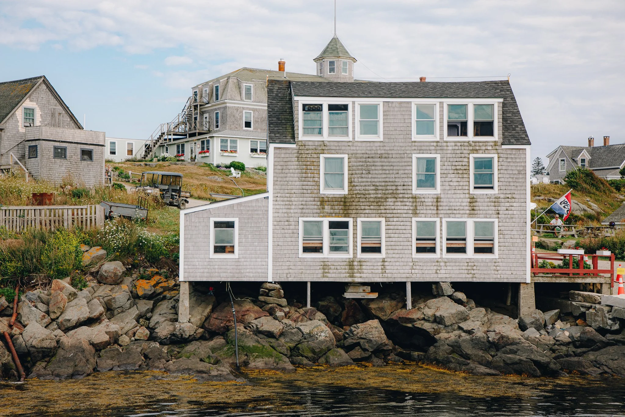House on stilts at the water's edge with rocks, grass, and a boat dock in the foreground, and other houses on a hill in the background under a partly cloudy sky.