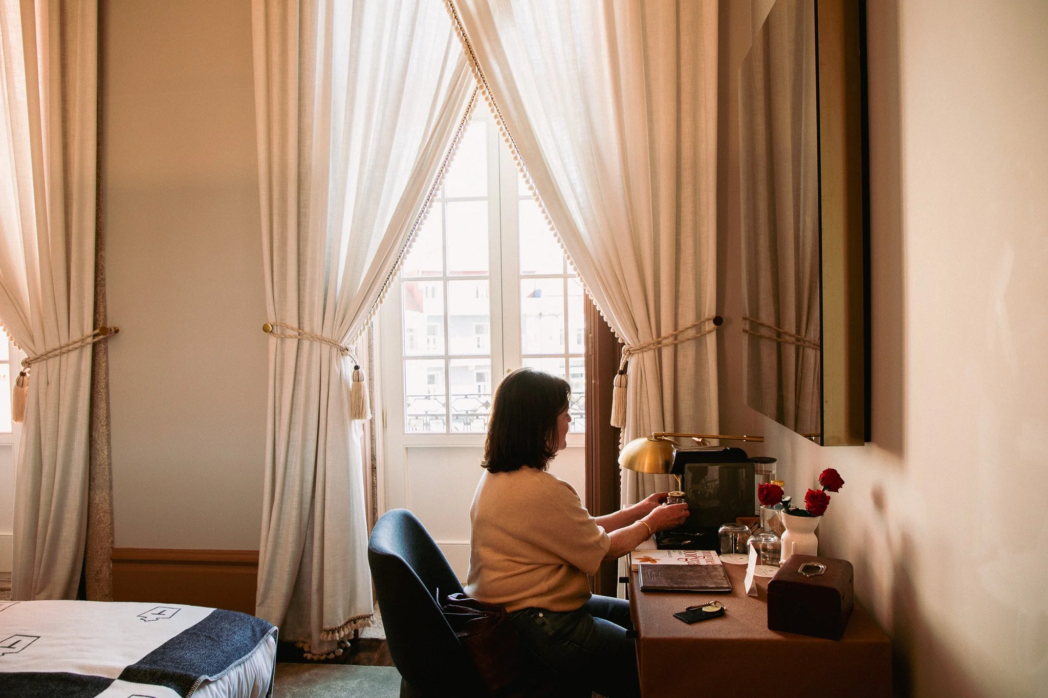 A woman sitting at a desk in a well-lit room with large windows, beige curtains, and a mirror on the wall, working on a laptop with a cup of coffee and a bouquet of red roses nearby.