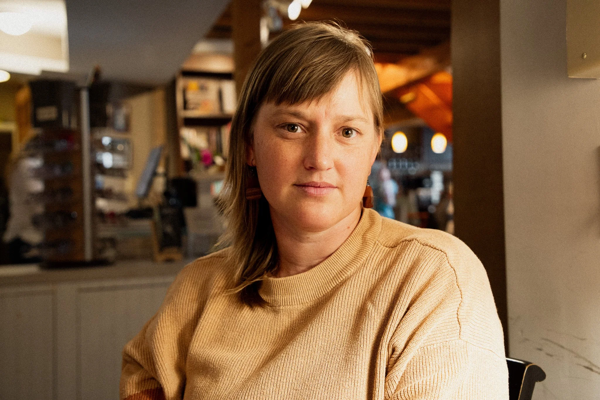 A woman with brown hair and light skin wearing a tan sweater, sitting indoors with a background of bookshelves and warm lighting.