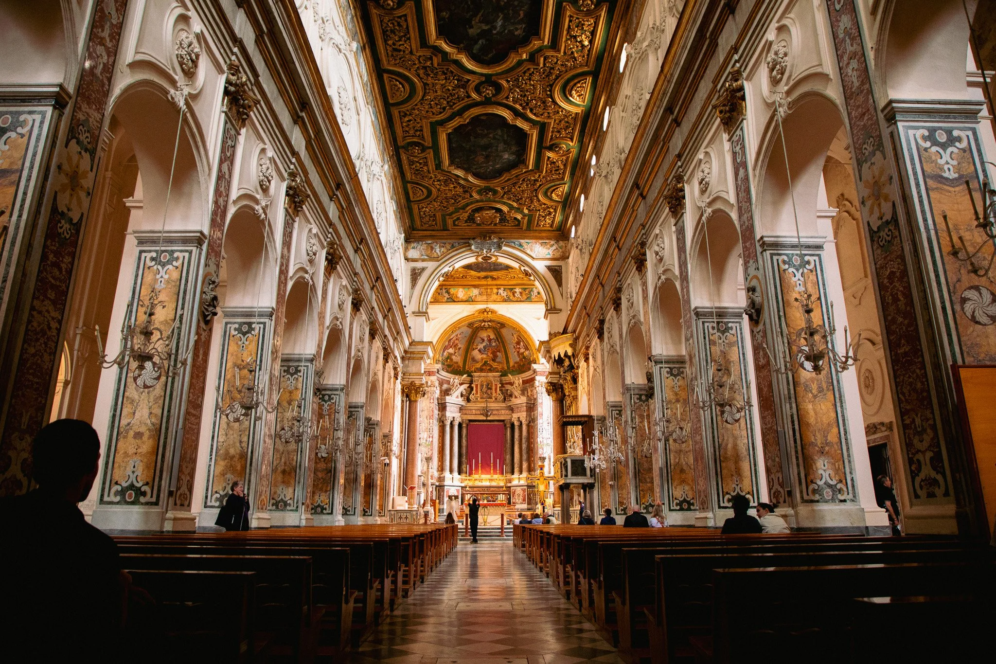 Interior of a grand, ornate church with high, decorated ceilings, statues, and stained glass windows, with several rows of wooden pews and people seated and standing.
