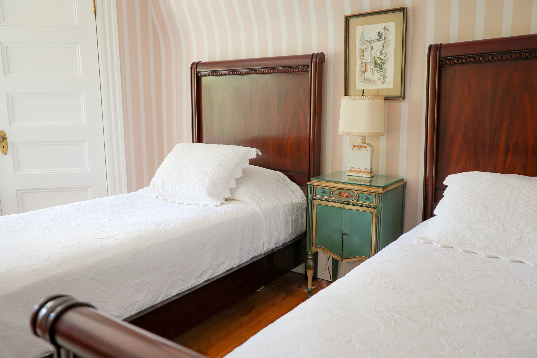 Two twin beds with white quilts and pillows in a bedroom with striped wallpaper. Between the beds is a vintage green nightstand with floral decoration, a table lamp, and a framed picture hanging above. A white door is visible to the left.