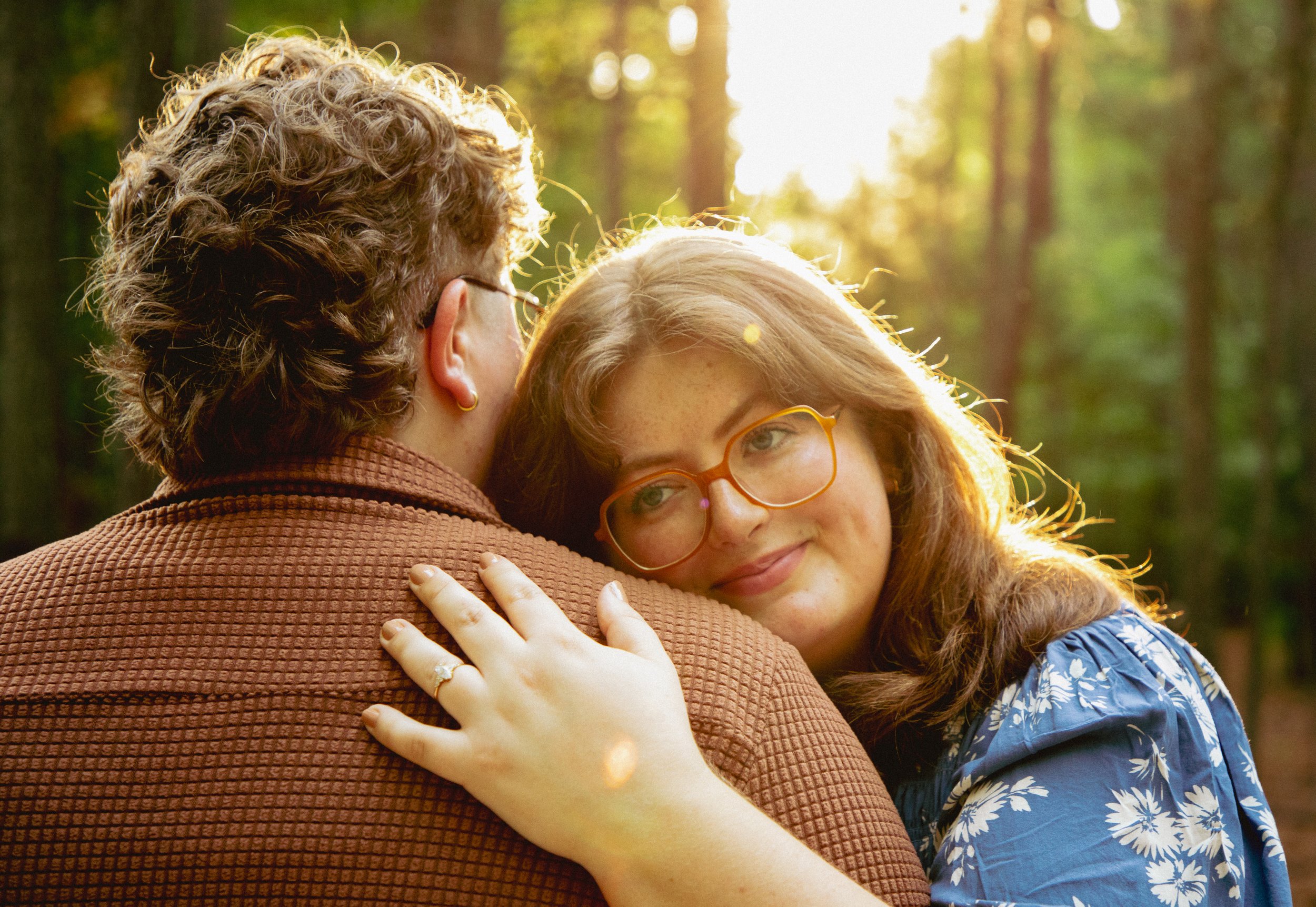 A woman with curly red hair and glasses hugging an older woman with curly hair, wearing glasses, in a forest with sunlight.