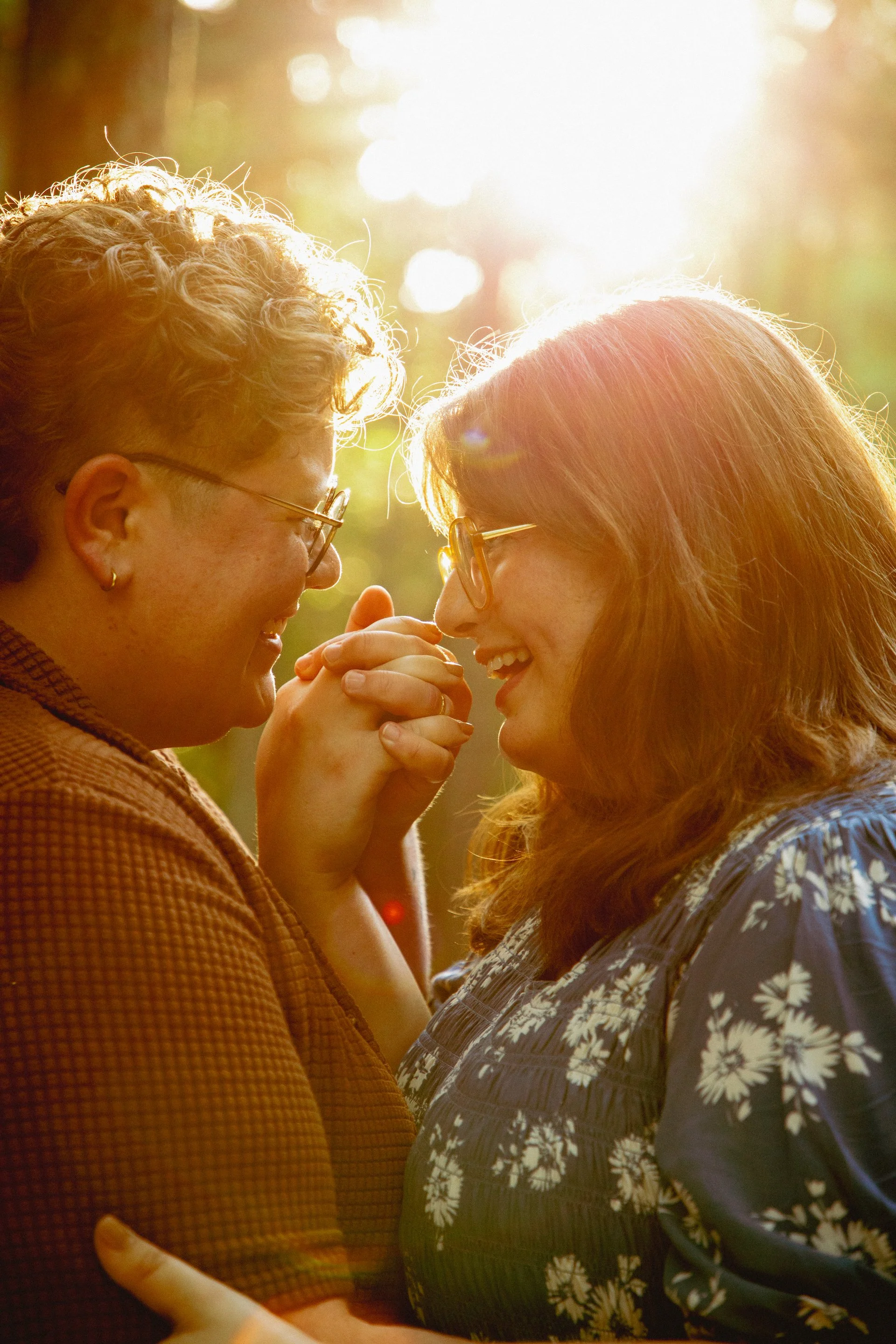 Two women face each other closely, holding hands, and smiling, with sunlight shining behind them in an outdoor setting.