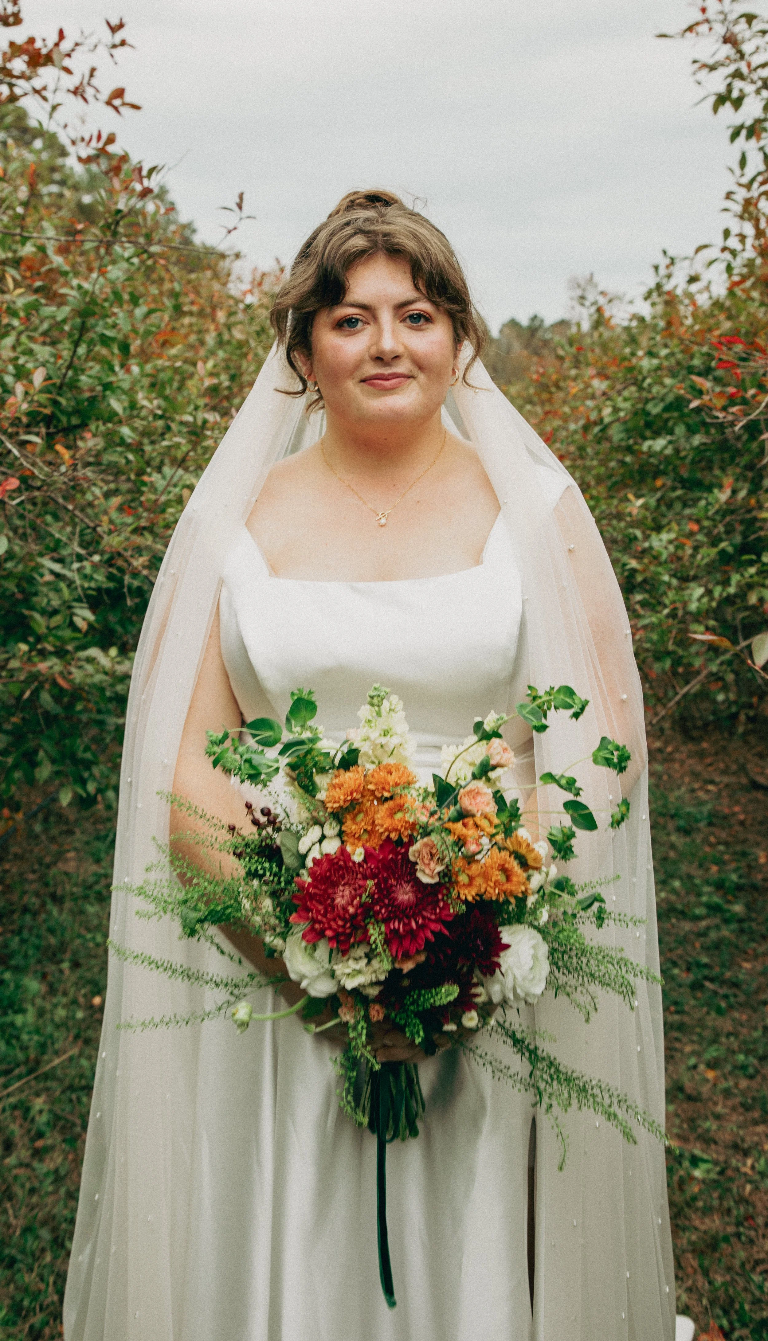 A woman dressed in a white wedding gown holding a colorful bouquet of flowers outdoors, with bushes and trees in the background.
