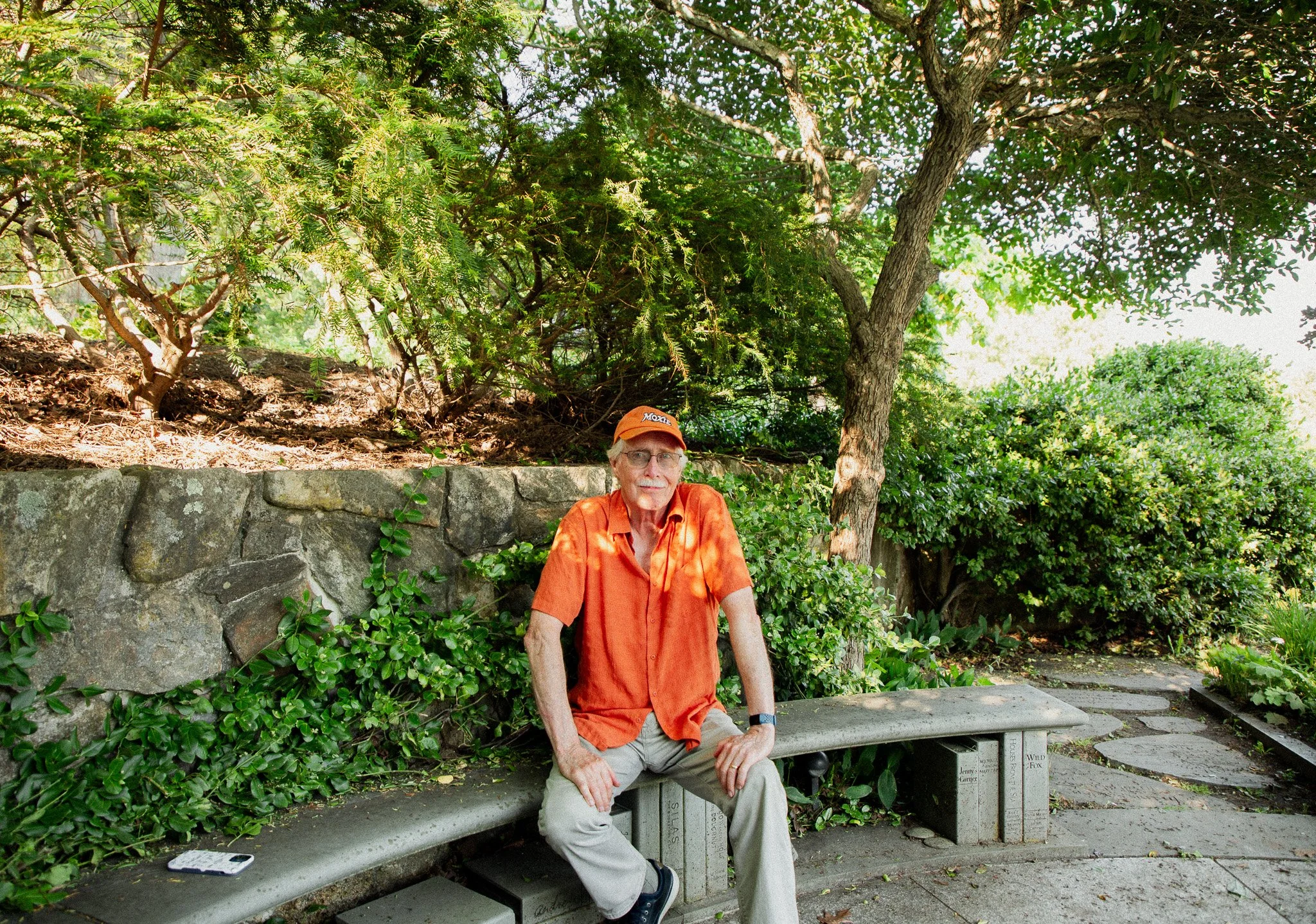 A man wearing an orange shirt and beige pants sitting on a stone park bench under green trees and bushes.