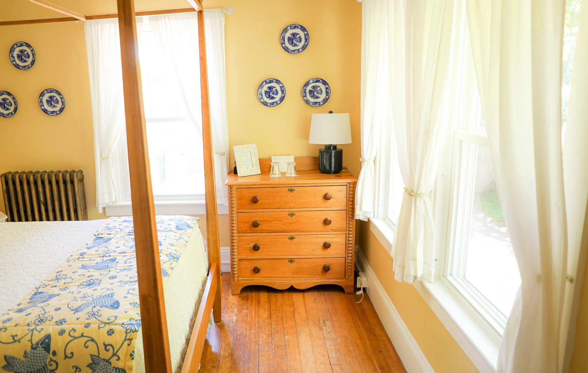 A sunlit bedroom corner with a wooden dresser, a table lamp, two glasses, framed paper, blue and white plates on yellow walls, white curtains, and a part of a bed with a patterned yellow and blue quilt.