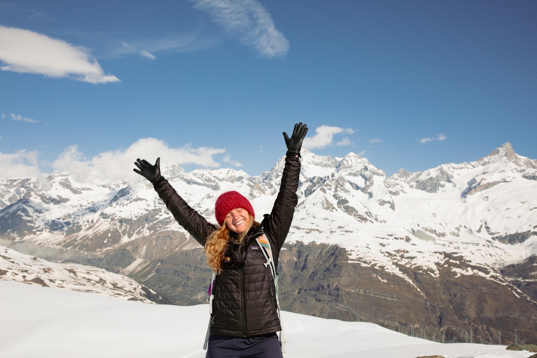 A woman with long curly hair, wearing a red beanie, black gloves, and a black jacket, smiling and raising her arms in the air, standing on snow-covered ground with snow-capped mountains and a blue sky with clouds in the background.