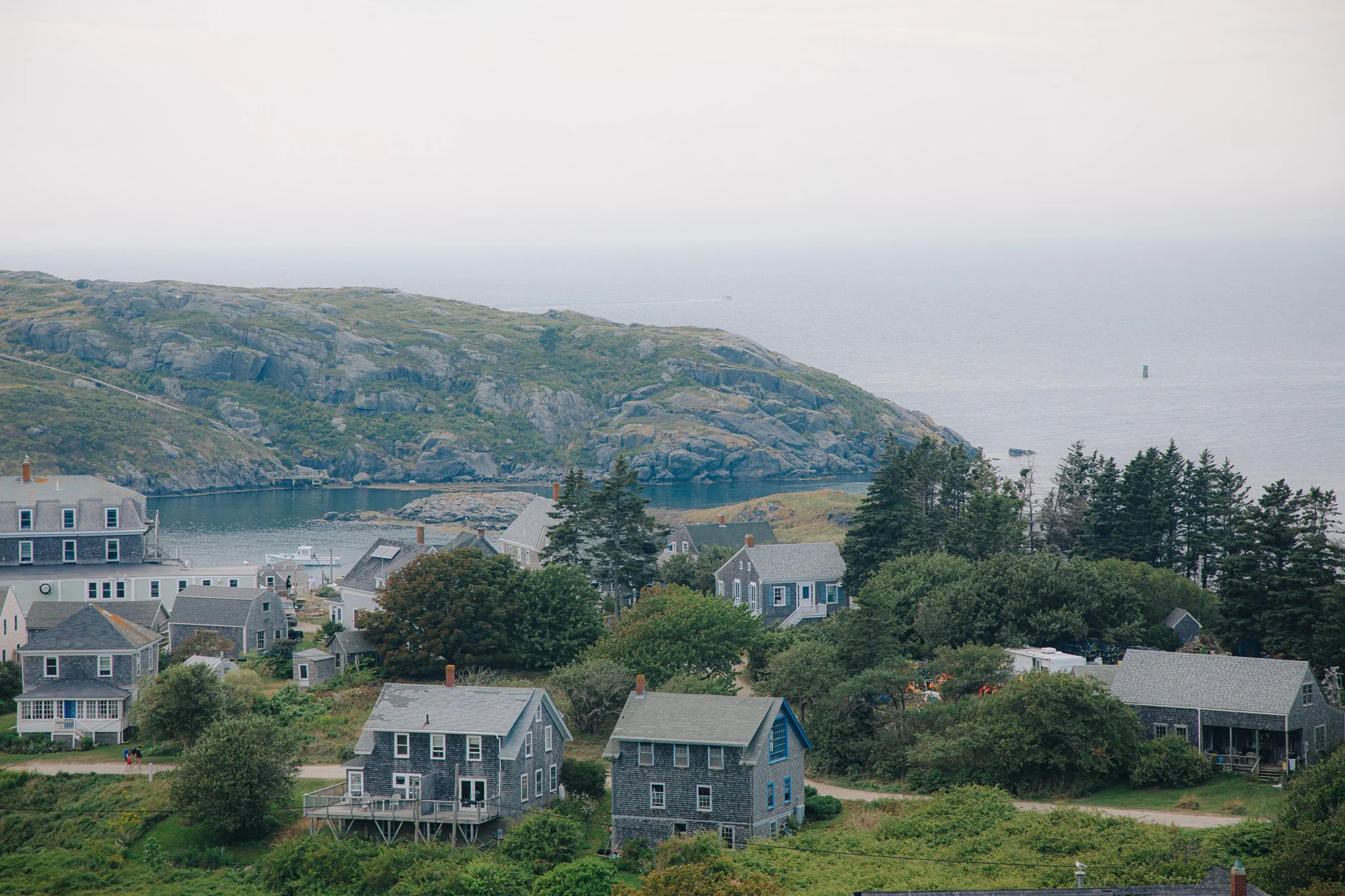 Coastal village with houses, trees, hillside, and ocean in the distance on a cloudy day.
