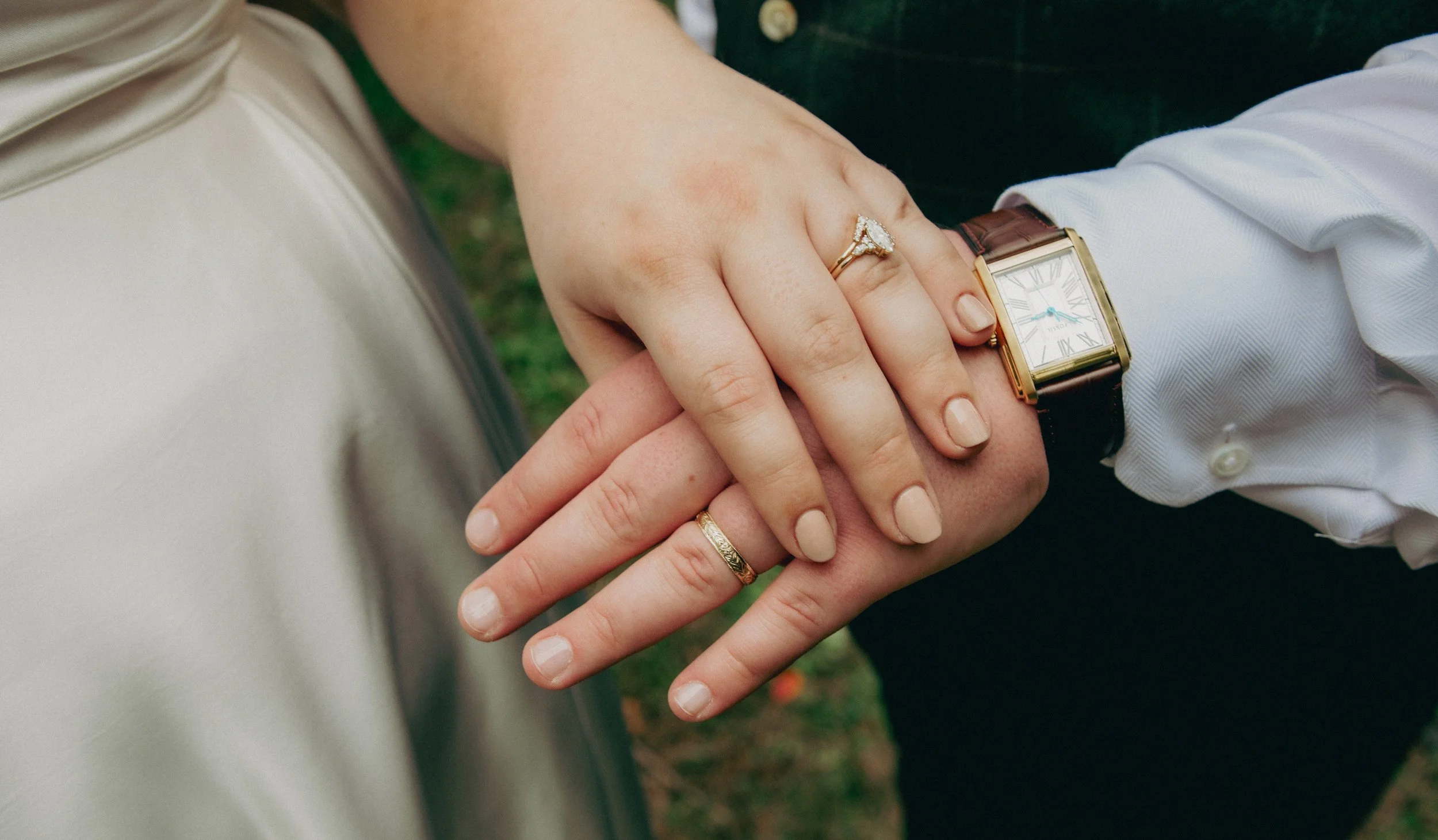 Close-up of a couple holding hands, with the woman's hand showing an engagement ring and wedding band, and the man's hand wearing a watch with a leather strap. The background is blurred greenery.