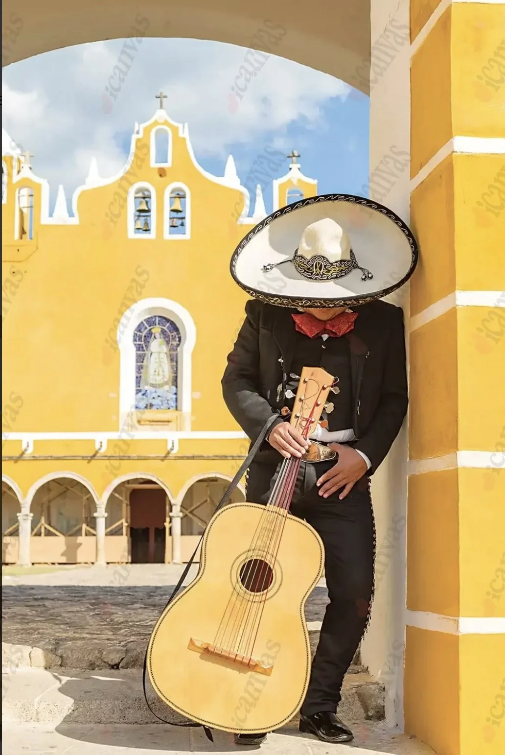 A person dressed as a mariachi musician, wearing a traditional sombrero and playing a guitar, stands next to a yellow colonial-style building with a bell tower and a religious image in the background.