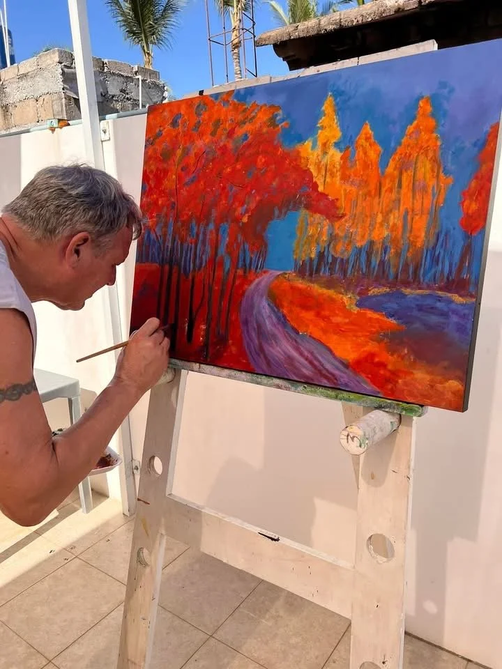 An artist painting an autumn landscape on a canvas. The scene features colorful orange, red, and yellow trees, a winding purple river, and a bright blue sky.