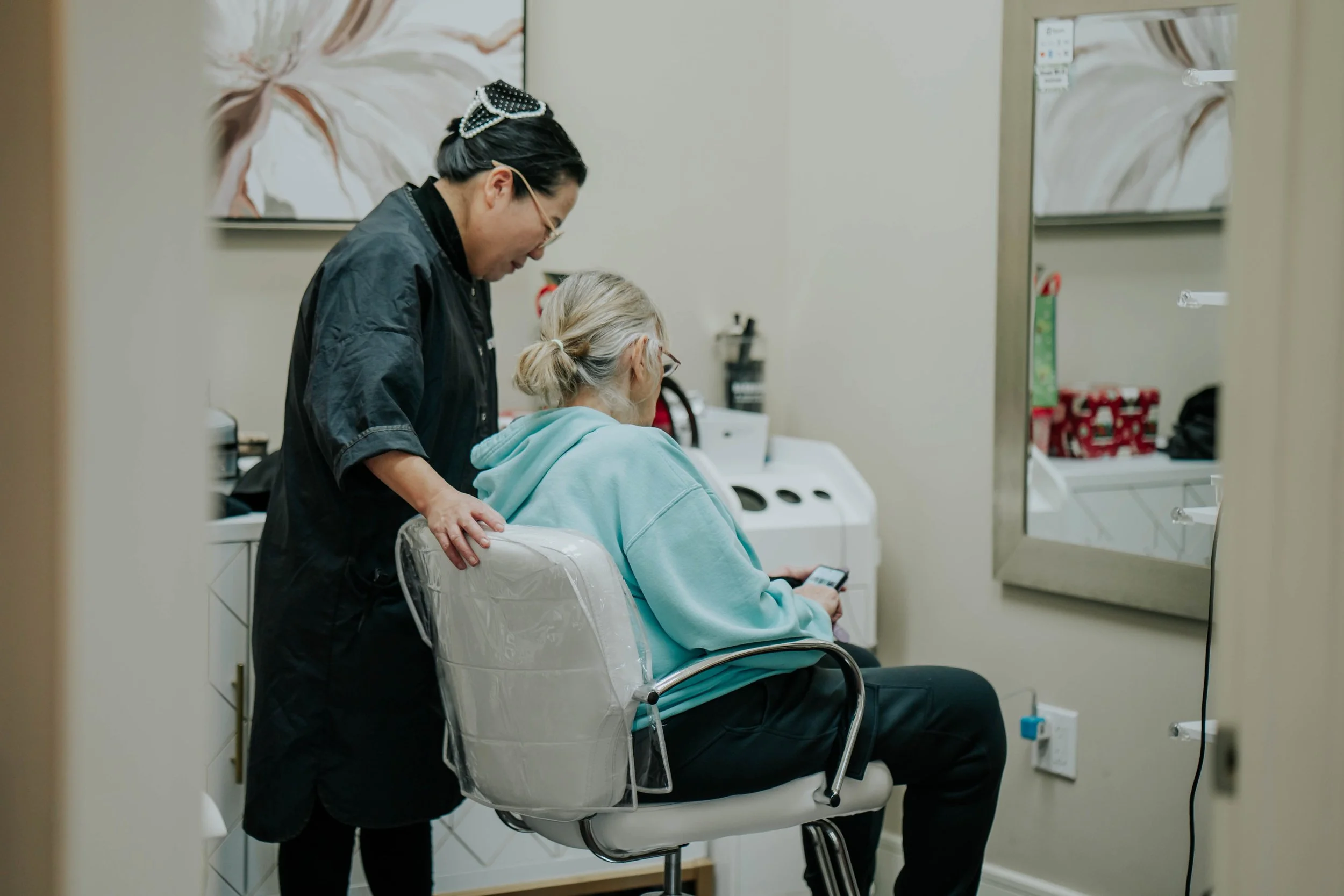 A healthcare worker in black scrubs shows a patient in a light blue hoodie something on her phone in a treatment room.