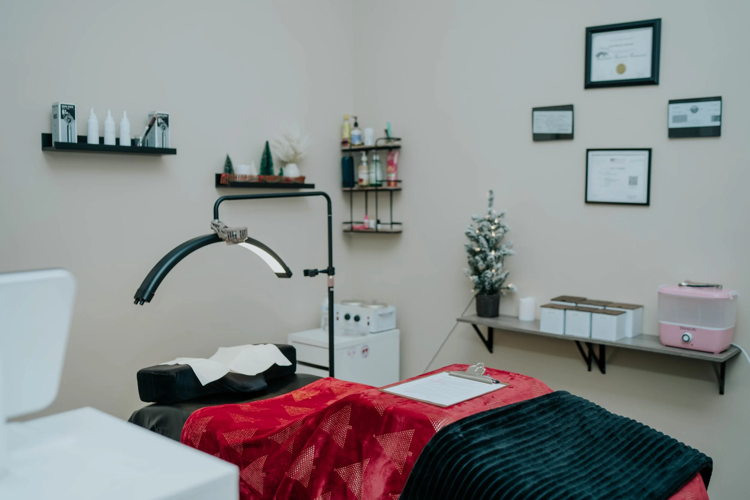 A cozy appointment room decorated with small Christmas trees and festive decor, featuring a treatment table with a red and black blanket, medical equipment, and framed certificates on the wall.
