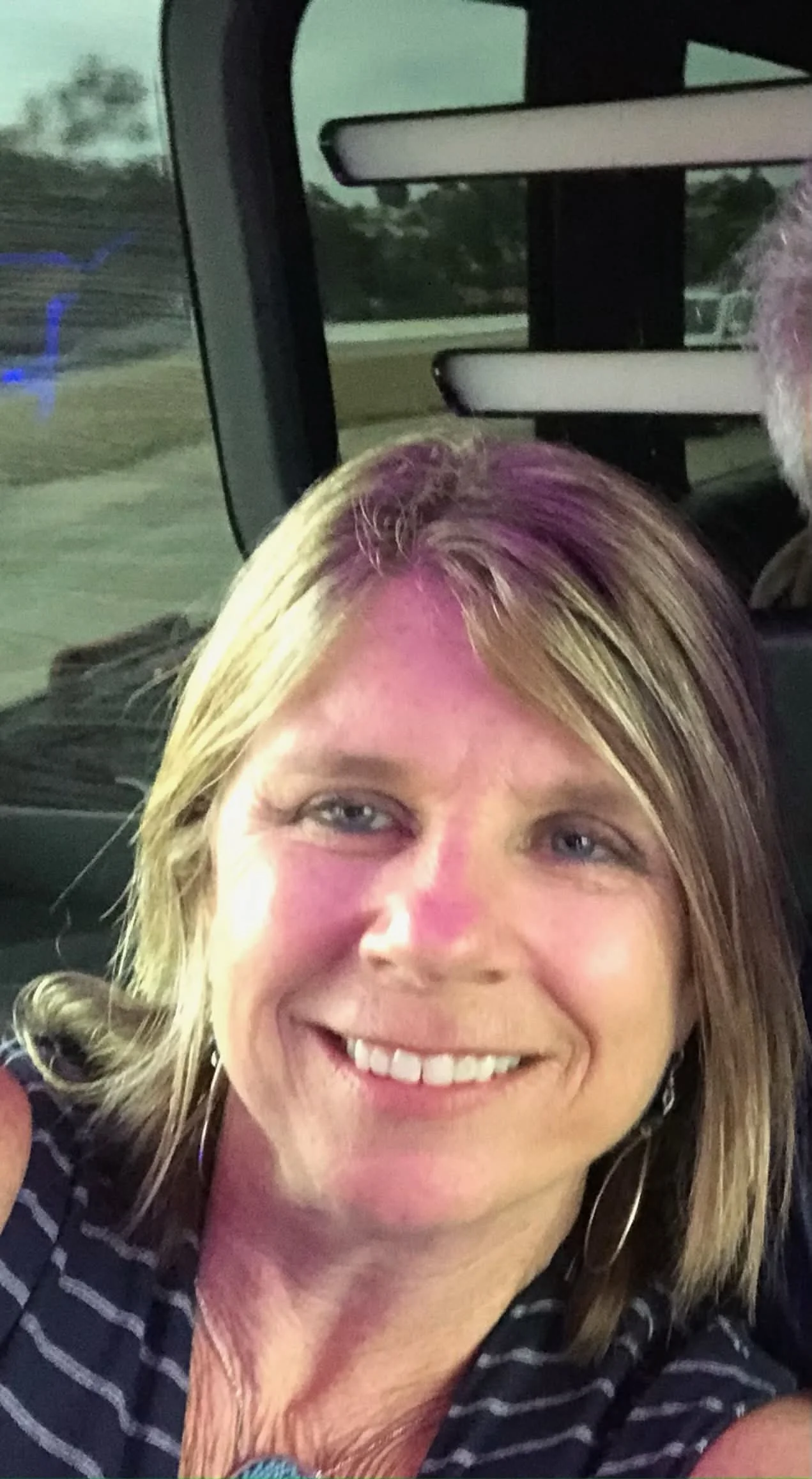 A woman with blonde hair and a striped shirt smiling inside a vehicle.