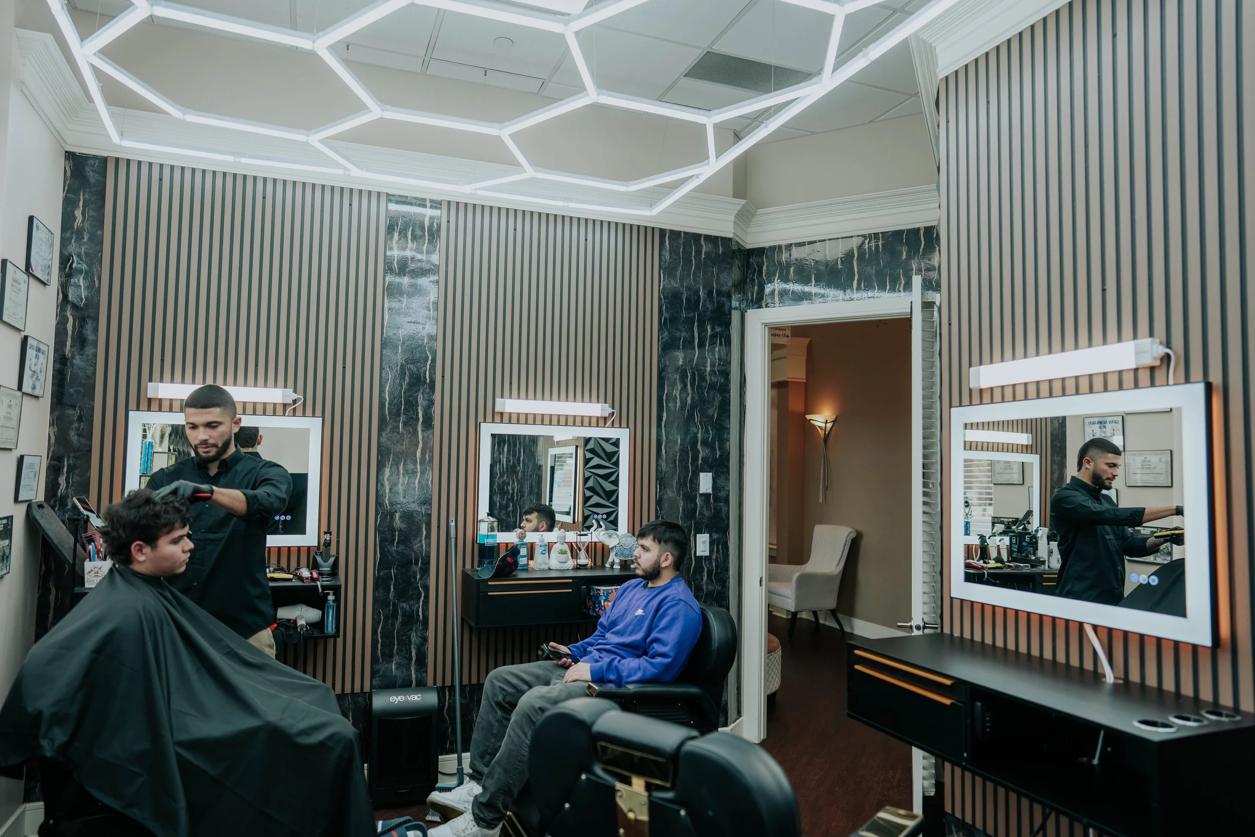 Inside a modern barbershop with two barbers and two clients, one client is getting a haircut while the other waits and looks at his phone. The room features striped walls, black marble accents, large mirrors, and a geometric ceiling light.