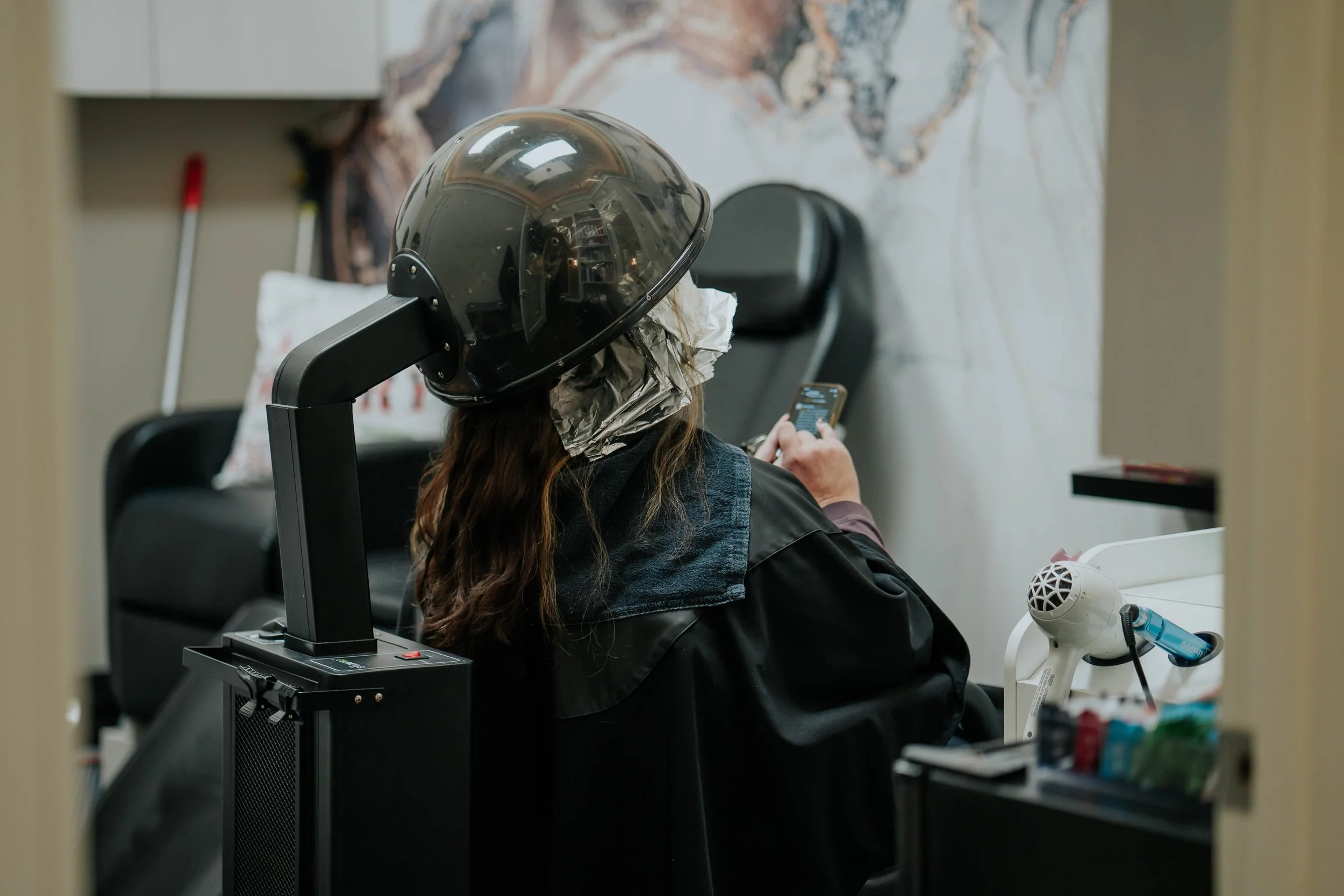 A woman sitting in a salon chair, wearing a hairdryer hood, looking at her phone. She is wearing a black cape and has brown hair. The salon has a hairdryer, a mirror, and hair styling tools nearby.