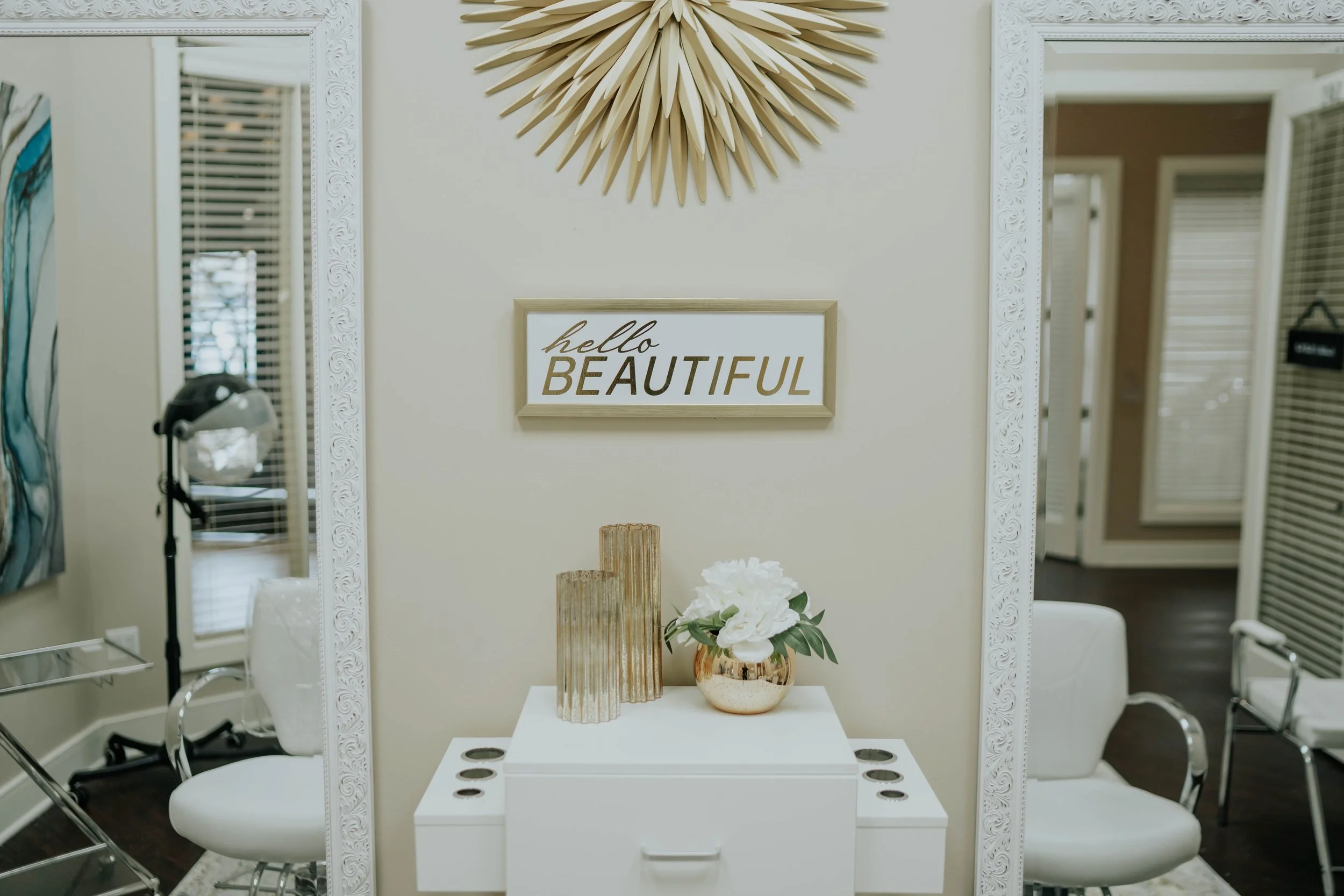 Decorative interior space with a framed sign that says 'hello BEAUTIFUL,' gold-colored vases, and a white flower arrangement on a white table. Mirrored wall panels reflect chairs and window blinds.
