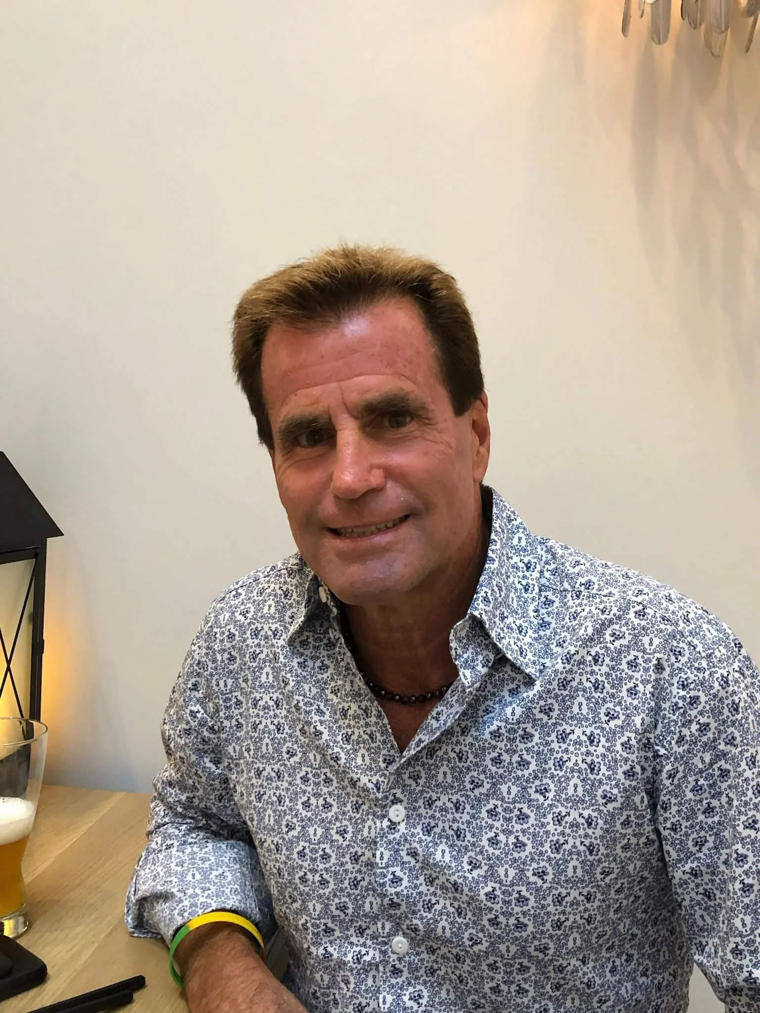 A man with short brown hair, wearing a white patterned shirt and a beaded necklace, sitting at a table with a glass of beer and a black phone.