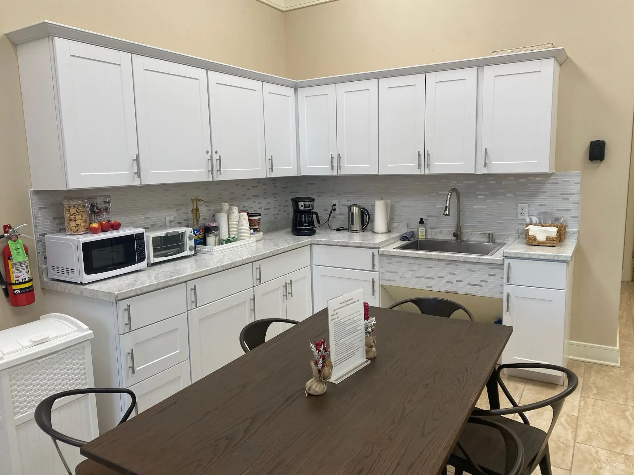 A kitchenette with white cabinets, a marble countertop, small appliances, and a dining table with chairs.