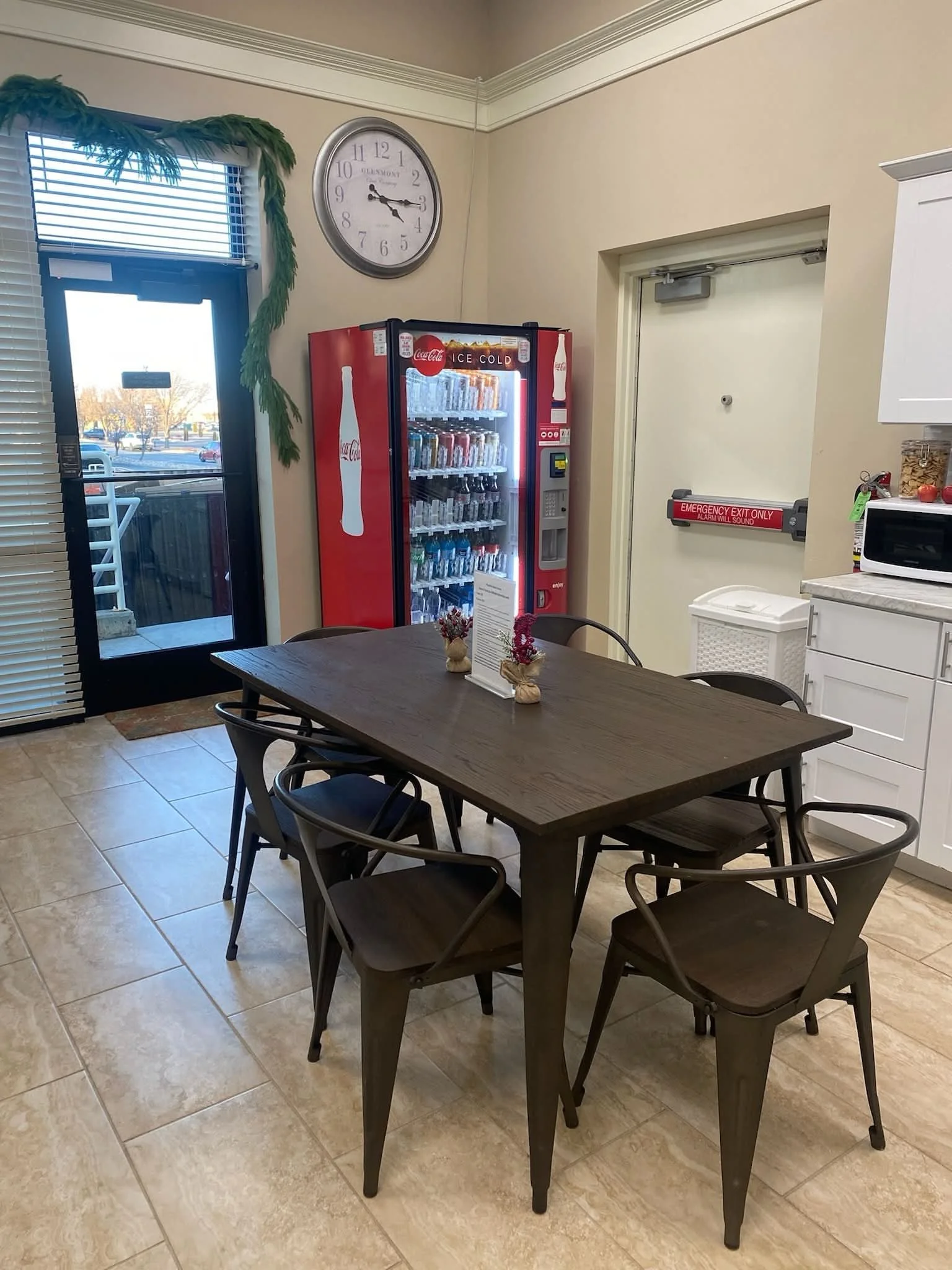 Interior of a small cafe or break room with a wooden table, six chairs, a soda vending machine, a clock, a door with an emergency exit sign, and a window