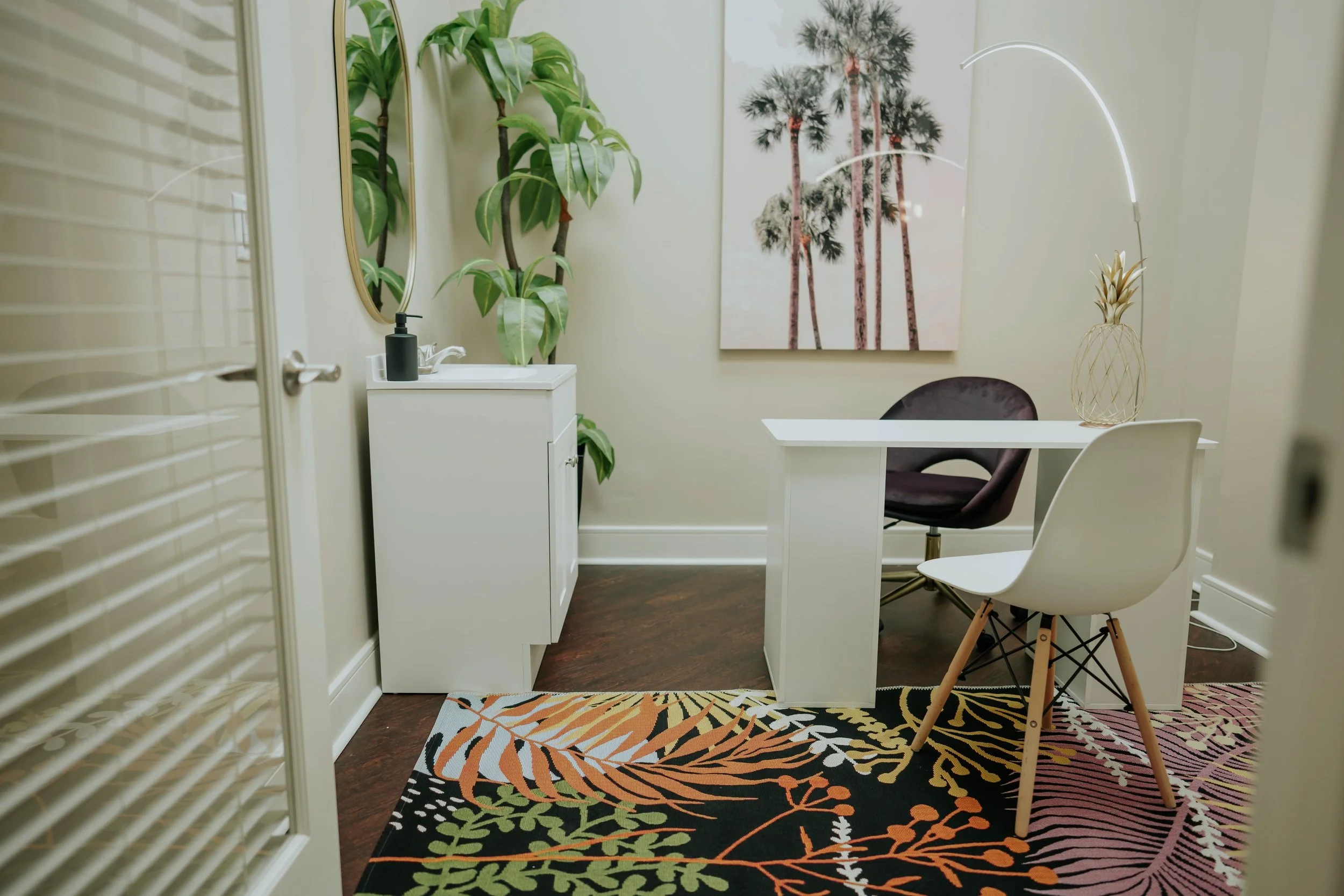 Small office with a white desk, brown and white chairs, tall indoor plants, framed artwork of palm trees, and a colorful tropical rug.