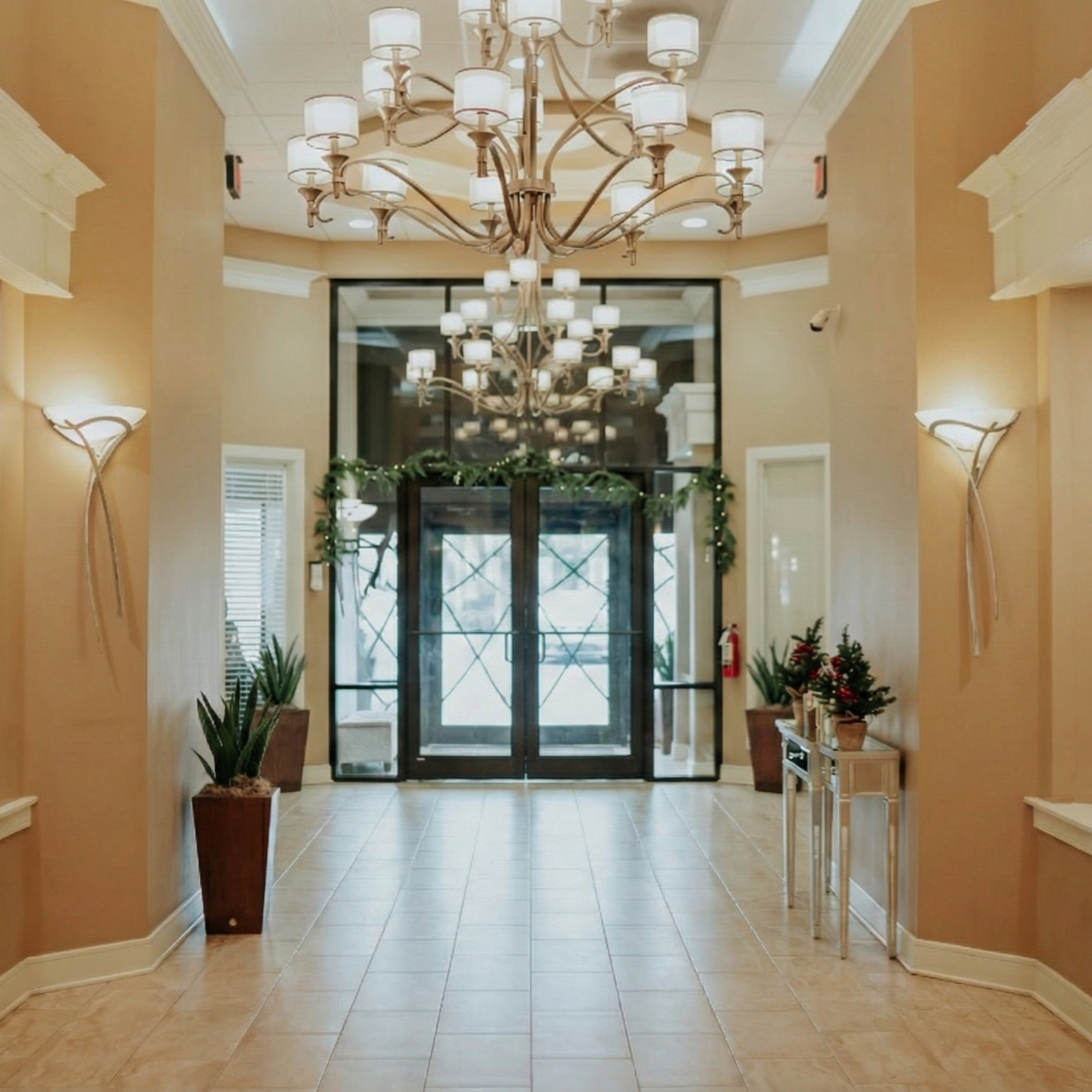 Elegant hotel lobby with beige walls, large chandeliers, wall sconces, potted plants, and a decorated table with a small Christmas tree.