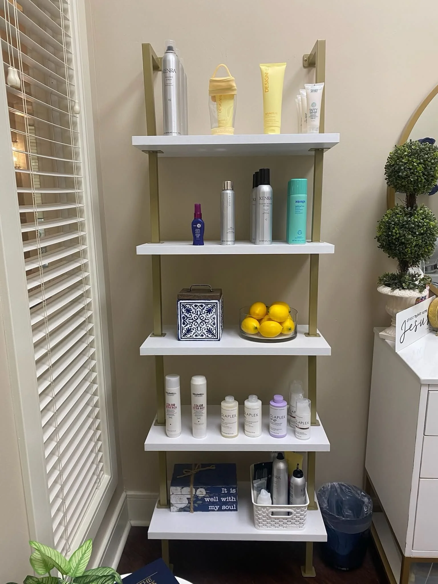 A gold and white shelving unit holding various hair and skincare products in bottles and tubes, a bowl of lemons, a decorative box, and a small basket, located in a room with a beige wall and nearby window blinds.