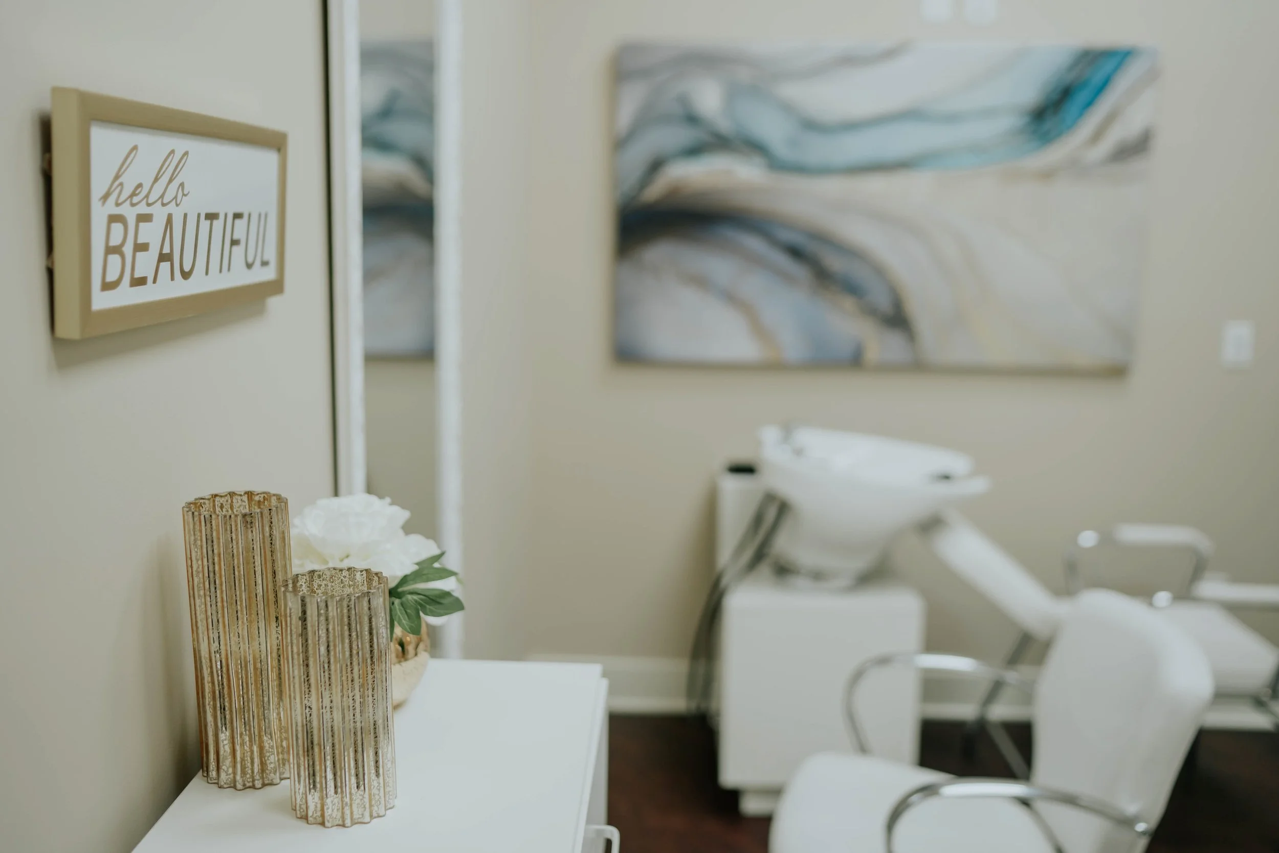 Decorative vases with flowers on a white table in a room with a beige wall, abstract wall art, and a sign reading 'hello beautiful'