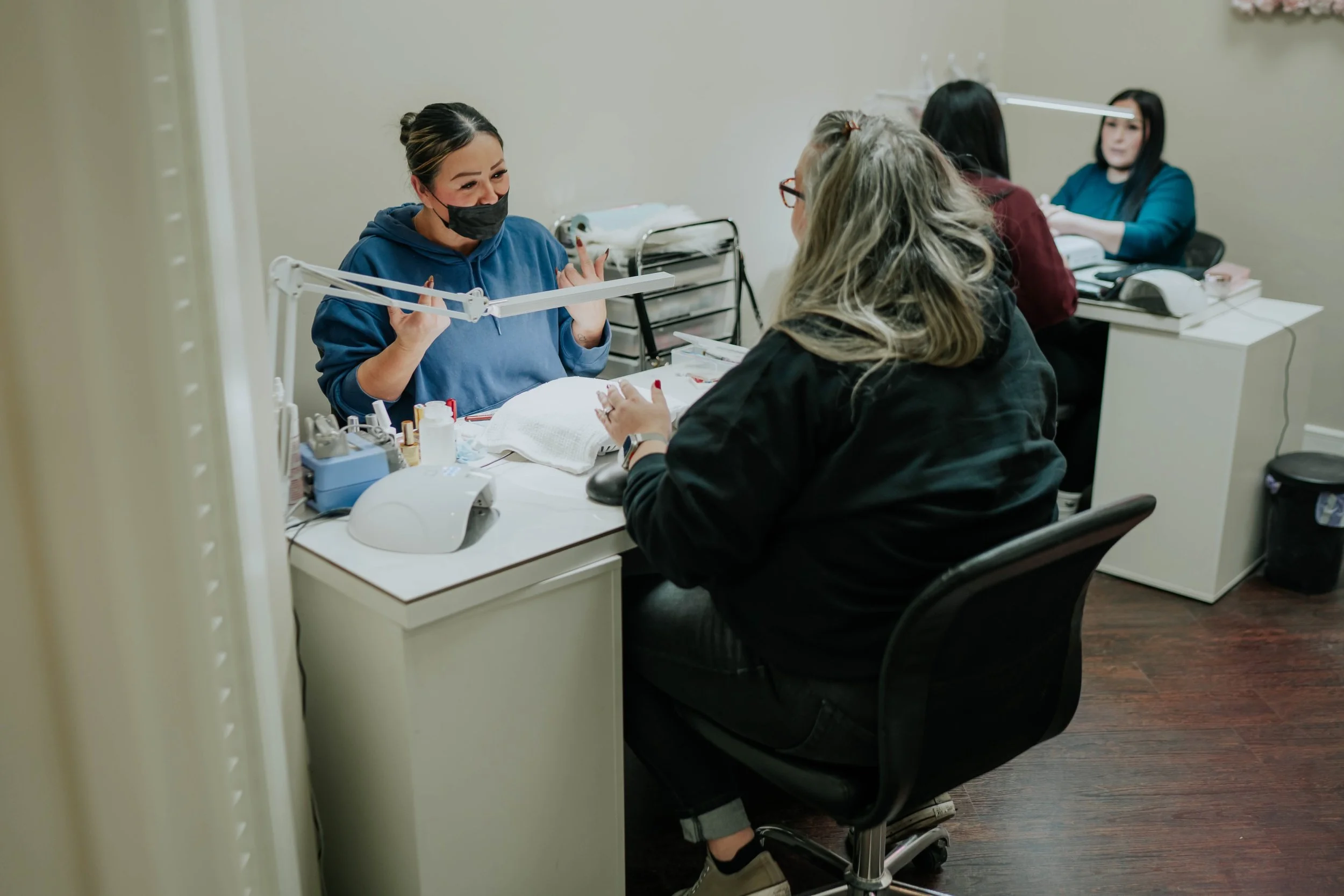 A woman getting a manicure at a nail salon, with two other women working on nails in the background.