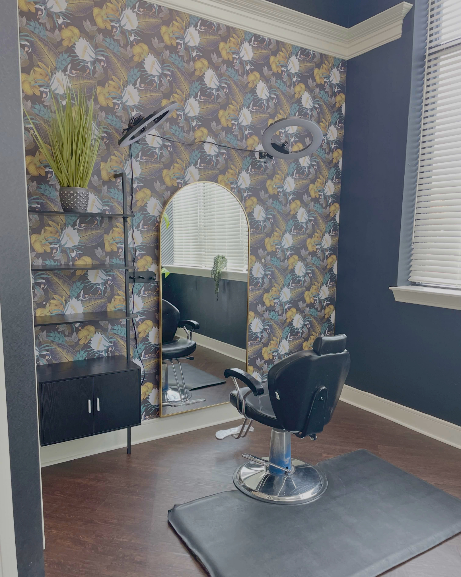 A hair salon station with a black styling chair in front of a large, arched mirror, a small black cabinet, a shelving unit with a potted plant, a ring light, wood flooring, wallpaper with floral pattern, and a window with blinds.