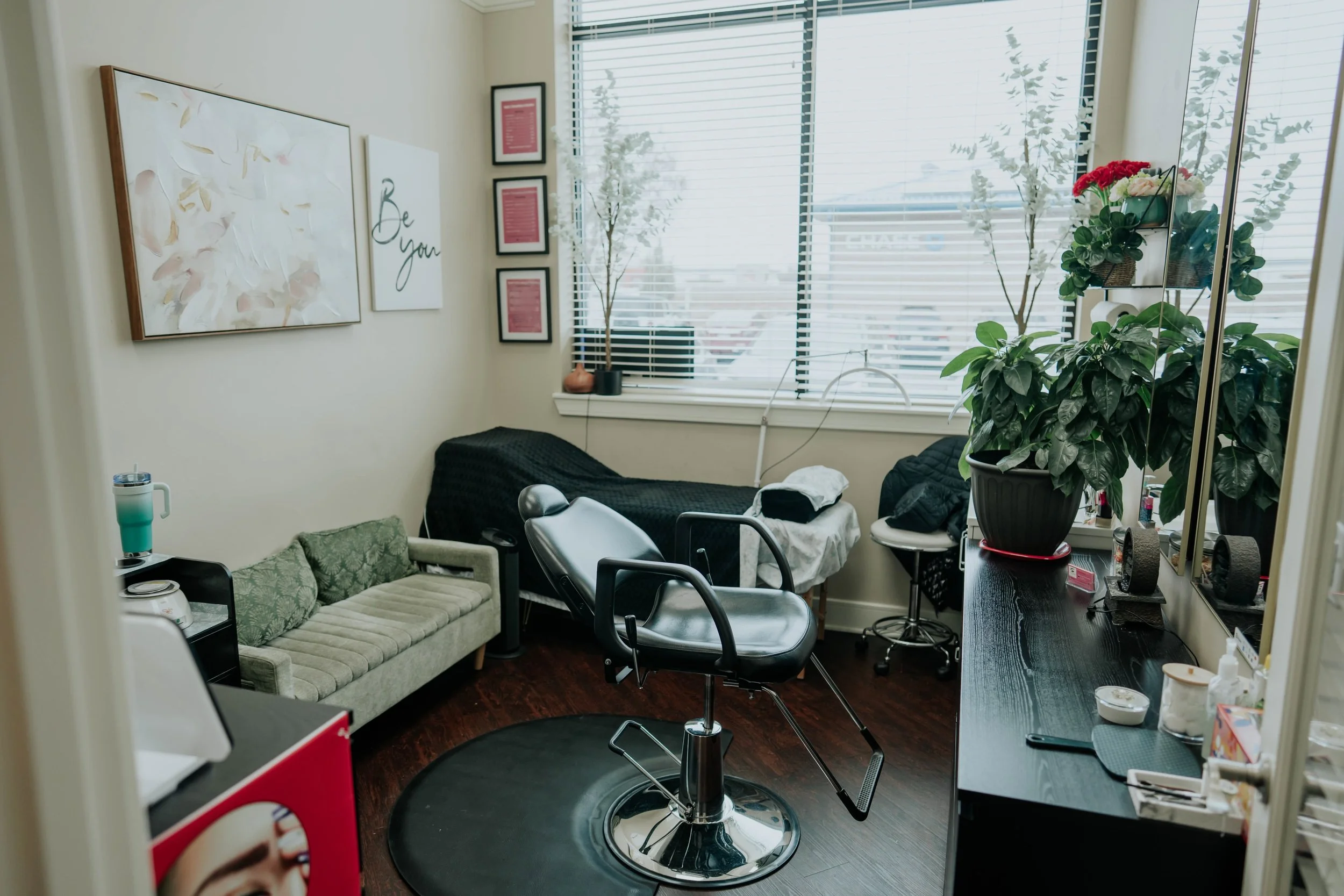 A small room with a black barber chair, a green couch, a white bed, and a large window with blinds. There are plants, framed artwork, a countertop with various items, and a large mirror.