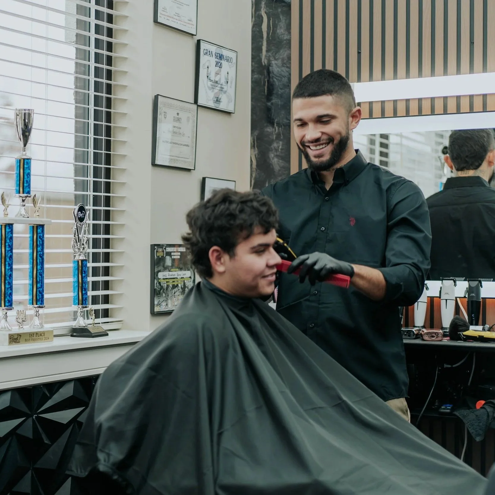 A man getting a haircut in a salon with trophies and framed certificates on the wall.