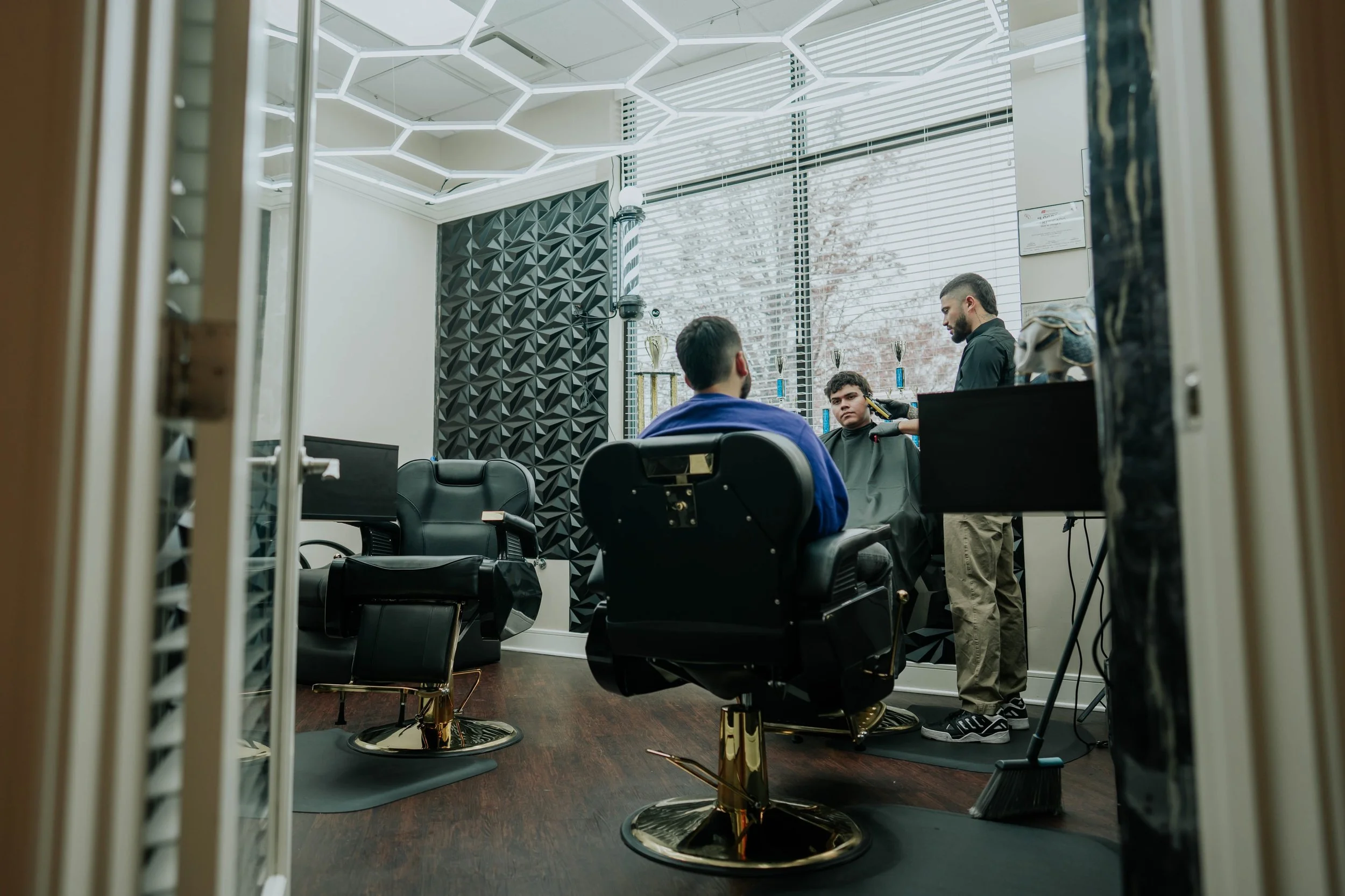 Two men are in a barber shop, one sitting in a barber chair and the other standing, both facing each other. The barber is using a clipper on the customer’s hair. The shop has modern decor, black leather chairs, and trophies on a shelf near a large window with blinds. Outside, trees are visible.