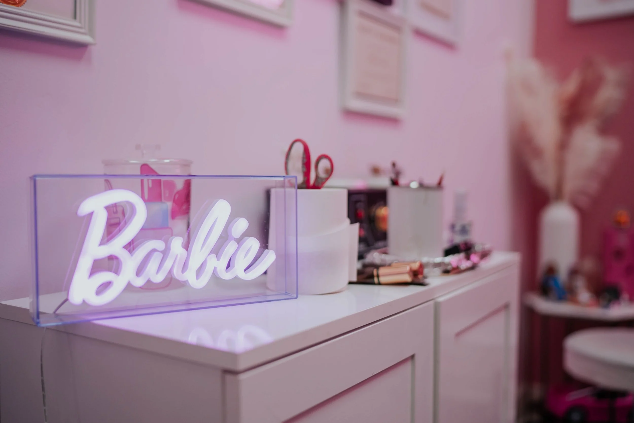 A pink-themed room with a white dresser holding various beauty and makeup items. In the foreground, a neon sign spelling "Barbie" sits inside a clear box, emitting a soft glow. The background features framed pictures on the pastel pink wall and decorative pink accents.