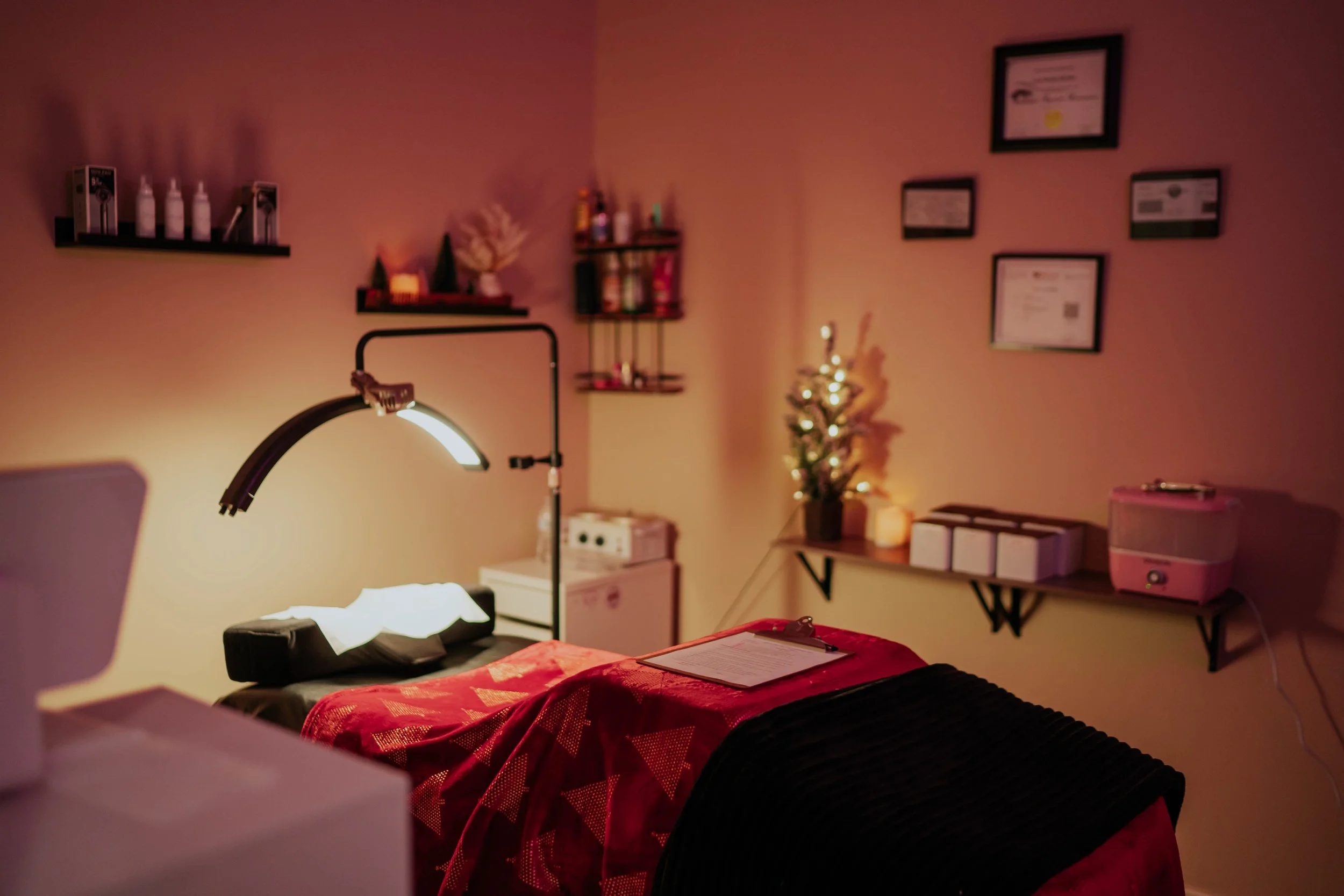 A massage therapy room decorated for Christmas with a small lit Christmas tree, candles, and framed certificates on the wall. A massage table with a red and black cover, a clipboard, and a lamp are in the foreground.