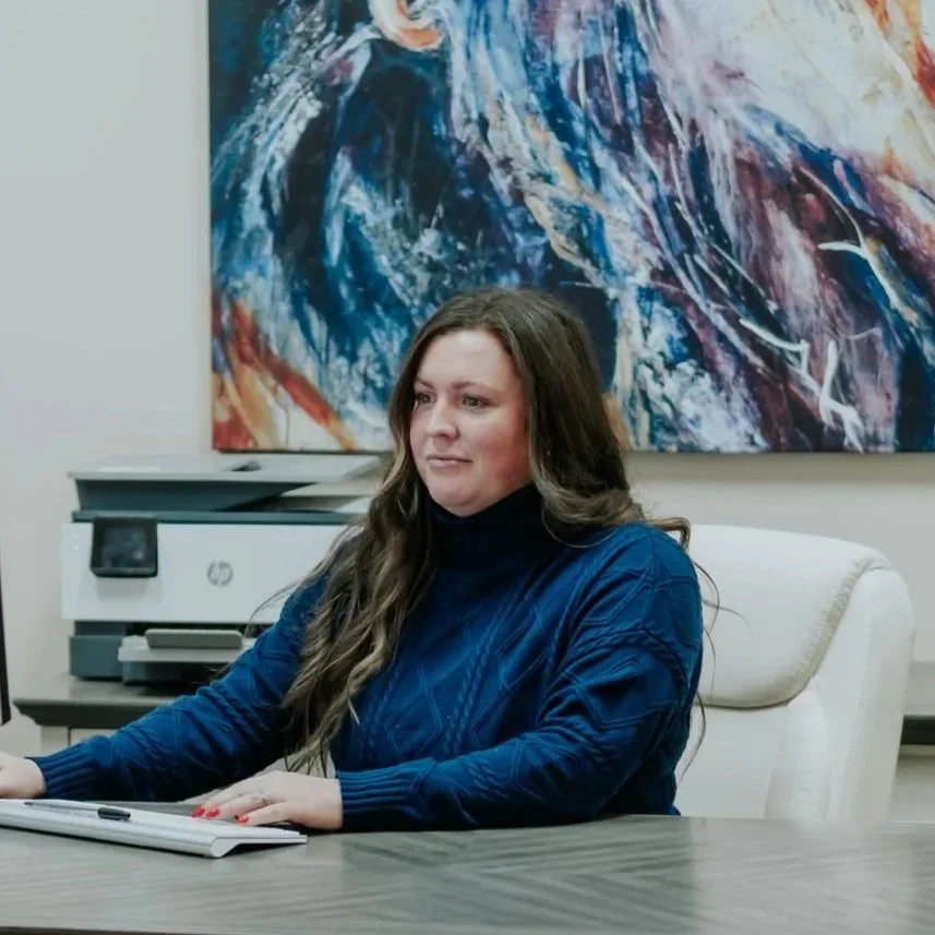 A woman with long brown hair sitting at a desk in an office, wearing a blue sweater, with abstract art on the wall behind her.