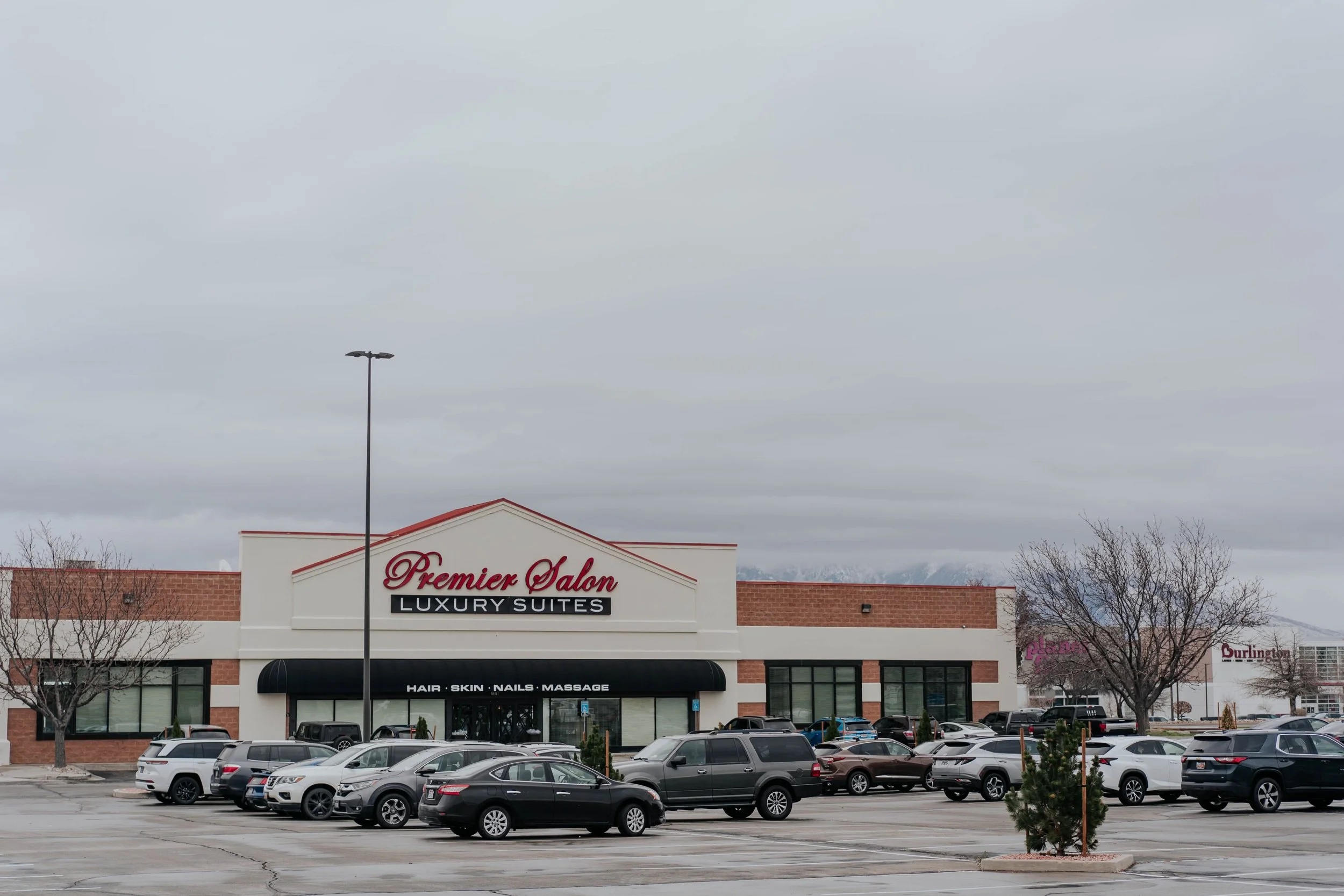 Parking lot in front of Premier Salon with luxury suites, hair, skin, nails, and massage services, in a shopping center with trees and parked cars, overcast sky.