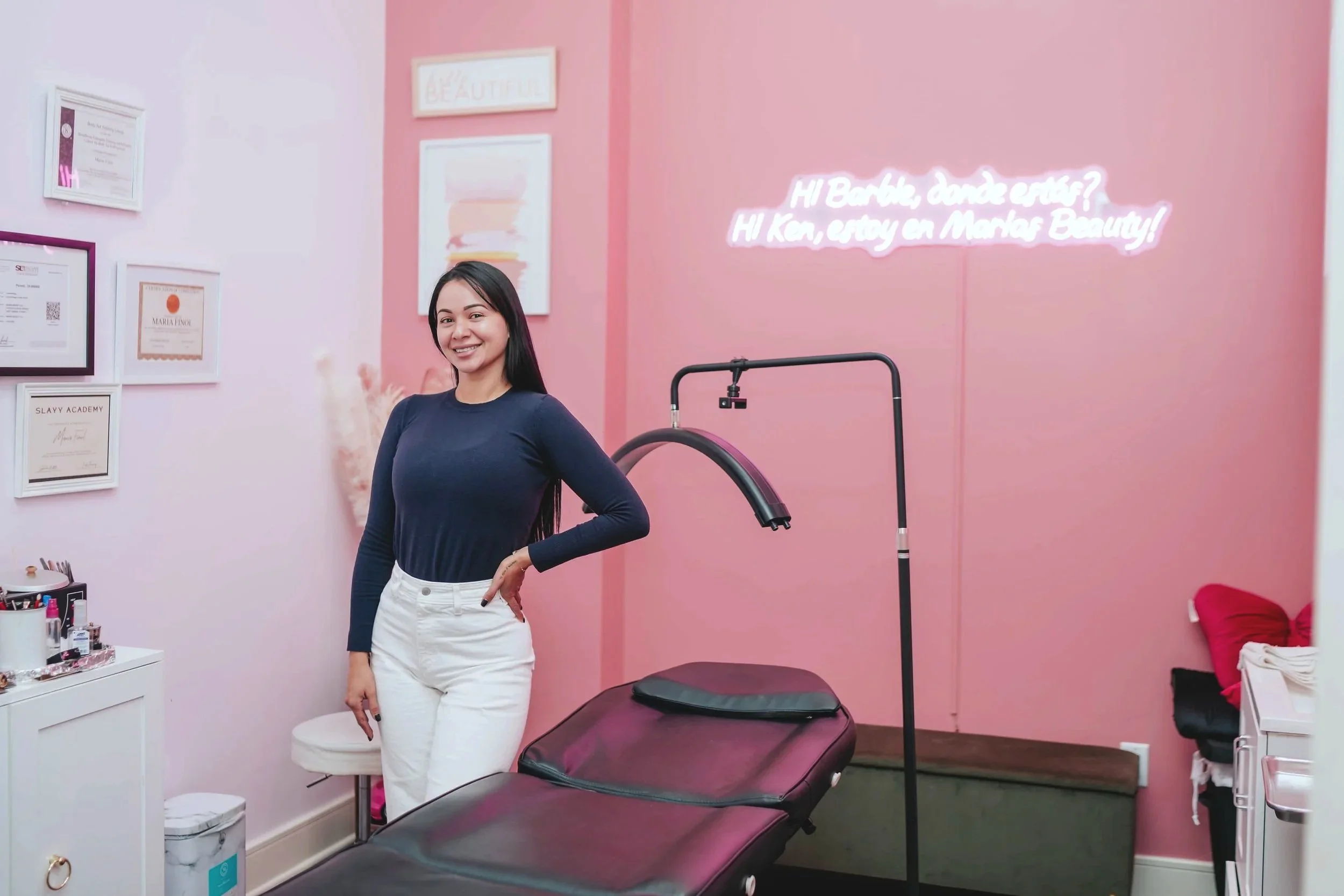 A smiling woman in a navy blue long sleeve shirt and white pants standing next to a treatment bed in a pink-themed beauty clinic room. There are framed certificates and artwork on the wall, a neon sign with a message, and a small white cabinet with beauty products.
