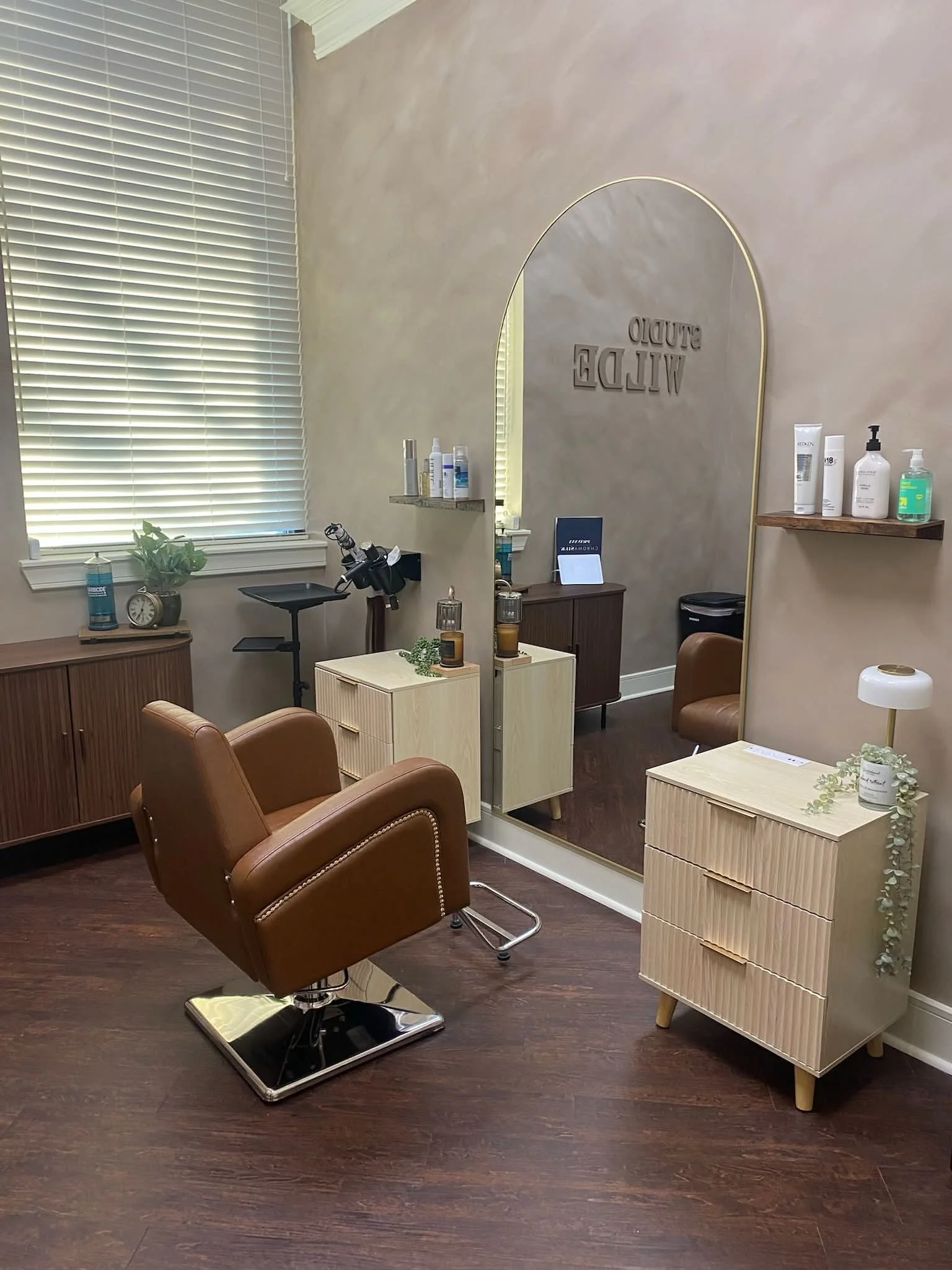 A cozy, well-organized hair salon setup featuring a brown leather salon chair, a mirror with an arched top, wooden cabinets, a small white dresser, and various hair care products on shelves and tables. The floor is dark wood, and there is a window with closed blinds allowing natural light to fill the room.