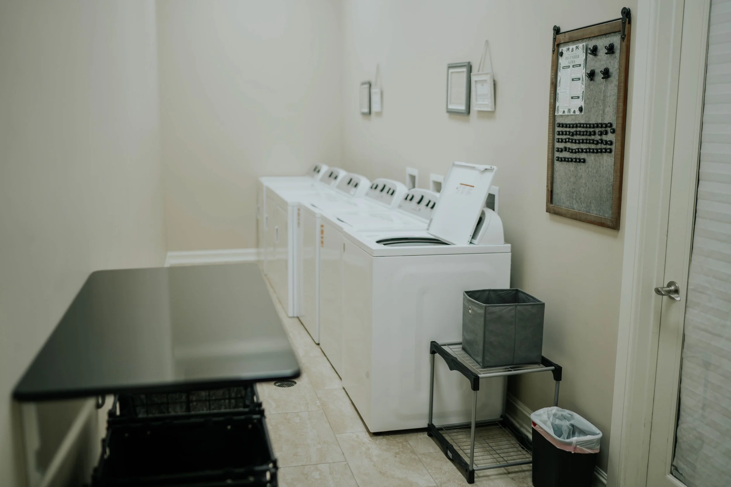 A row of white laundry washing machines in a laundry room with framed pictures hung on the wall above and a notice board with hooks to the right.