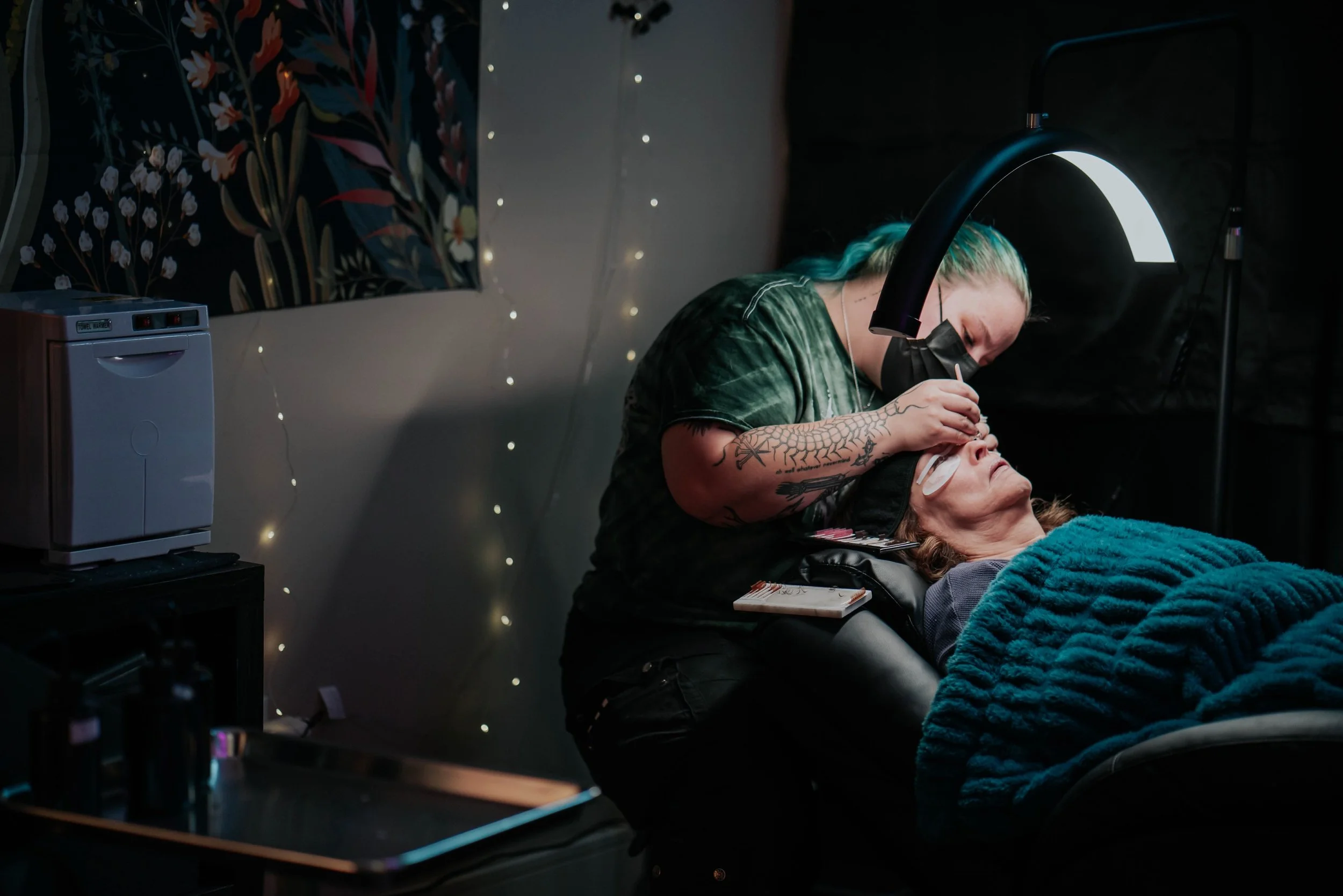 A woman receiving eyelash extensions from a technician in a dimly lit salon with fairy lights and a floral wall hanging.