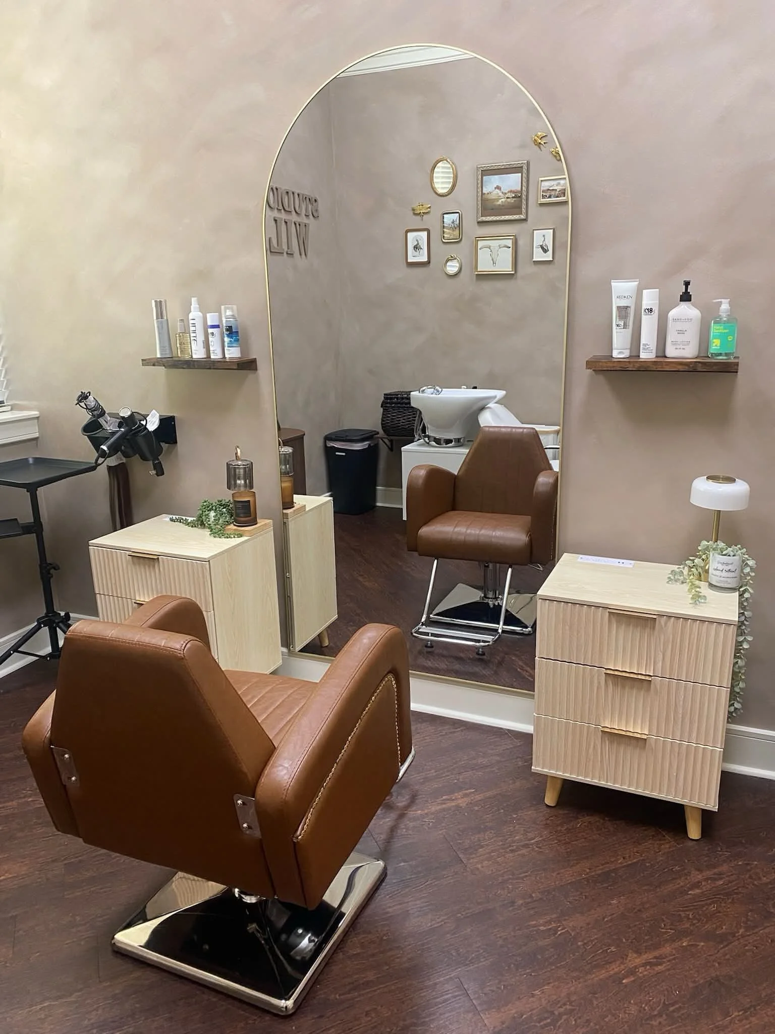 An interior view of a hair salon or barber shop with a styling chair, a small side table, a mirror, and a washing station in the background, decorated with framed pictures and wall decor.
