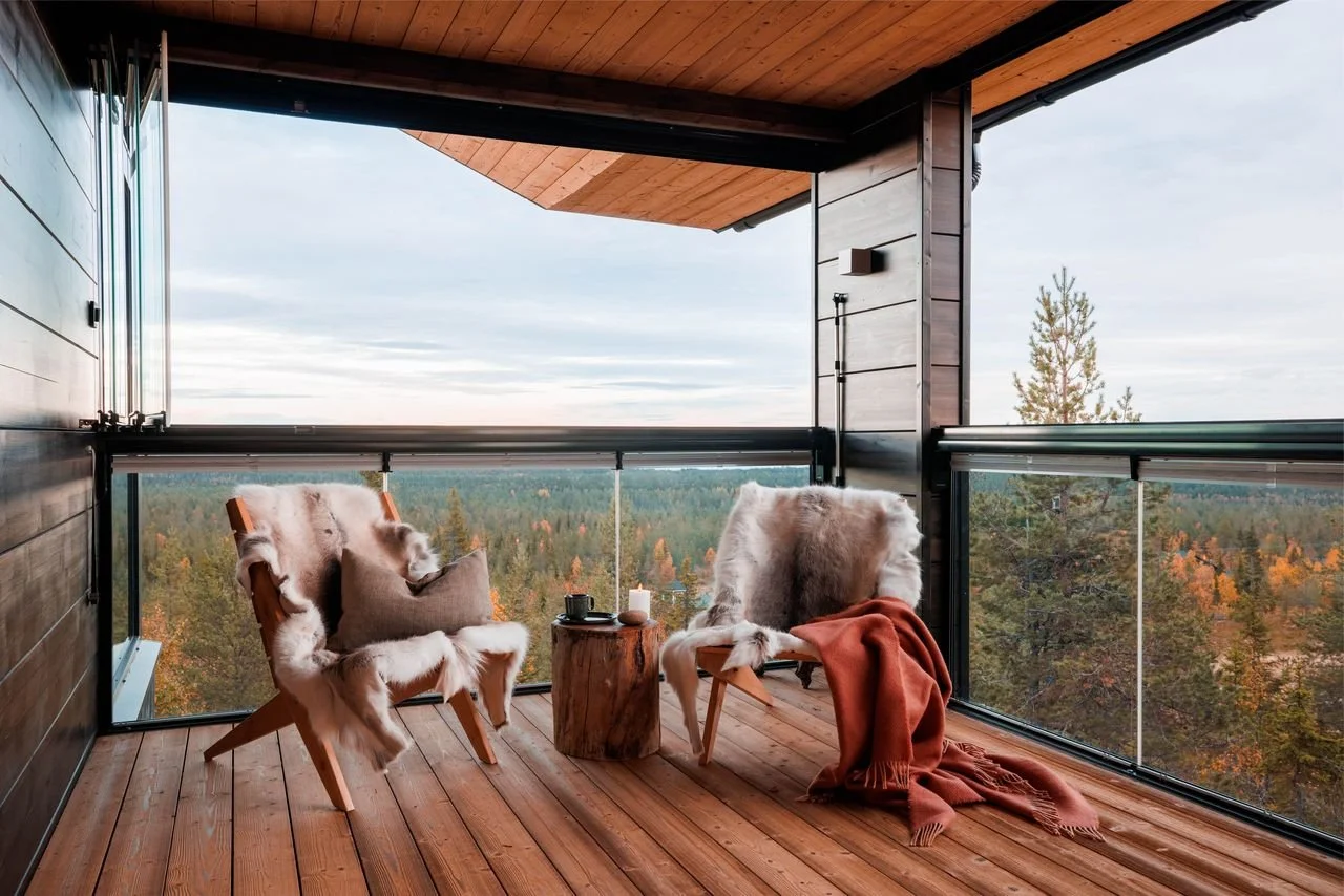 A cozy balcony with two wooden chairs draped in fur throws, a small tree stump table holding cups and candles, overlooking a scenic forest view with trees displaying fall colors.