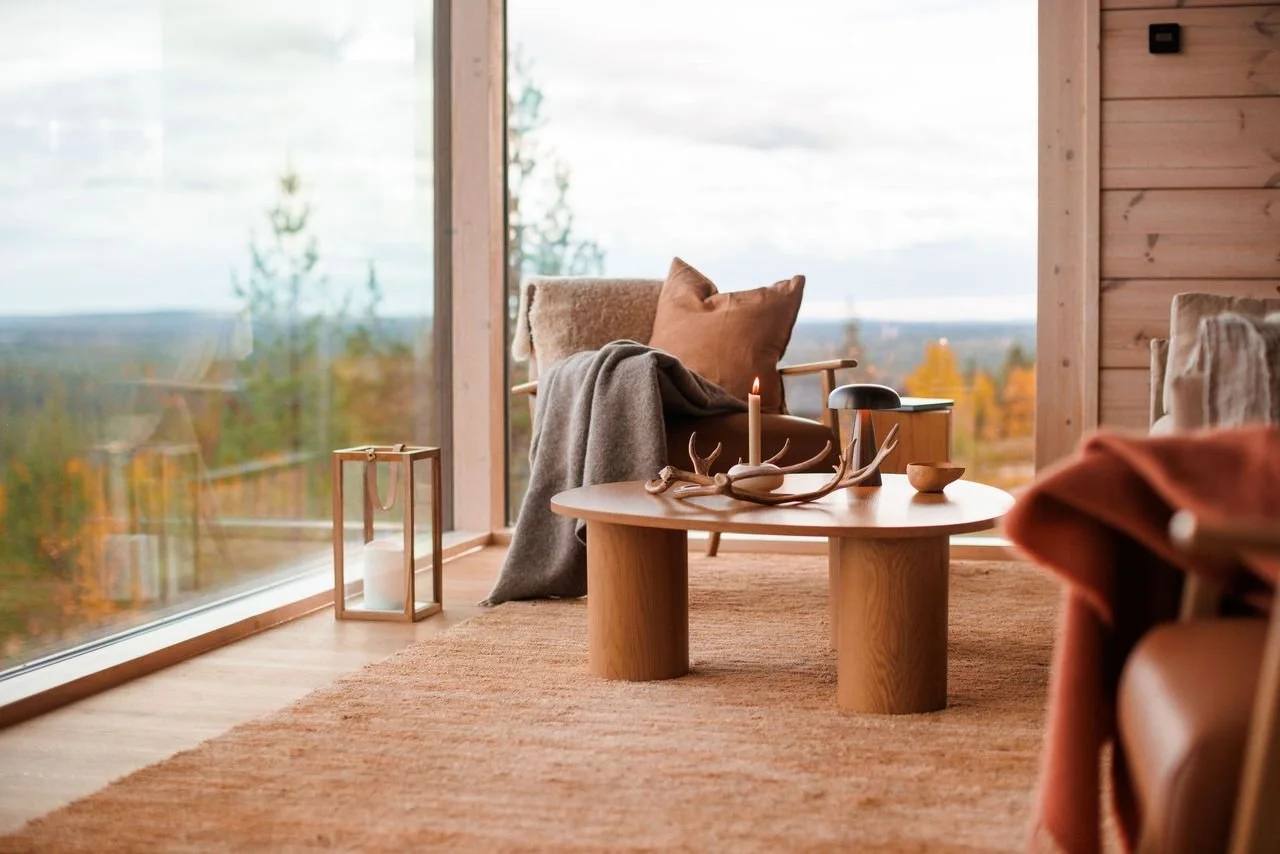 Cozy living room with wooden walls, large window overlooking trees and sky, round wooden coffee table with candles and decorative antlers, beige armchair with cushions and blanket, lantern with candle, and warm earthy tones.