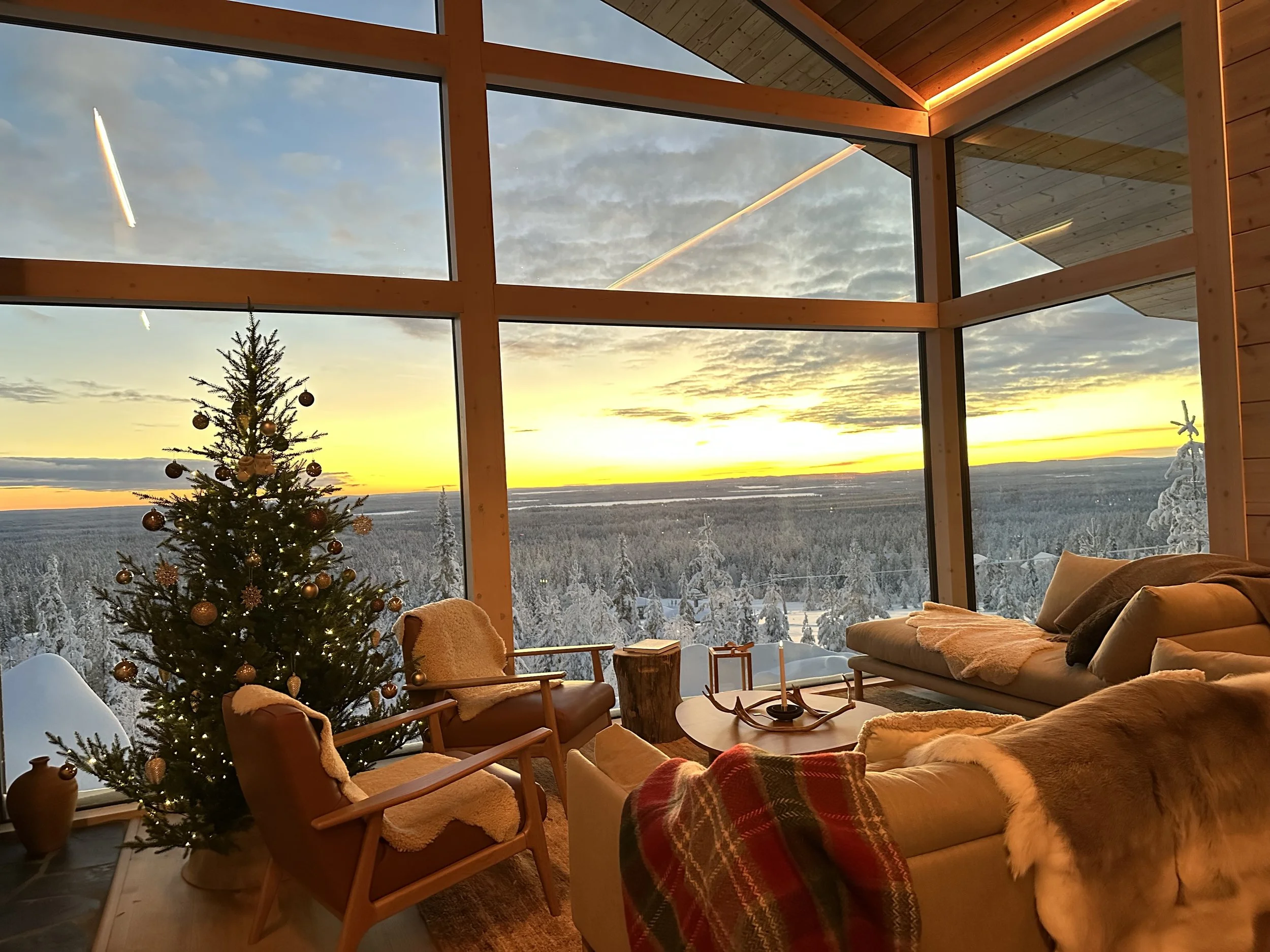 Living room with large floor-to-ceiling windows overlooking a snowy landscape at sunset, decorated Christmas tree, comfortable chairs, and cozy blankets.